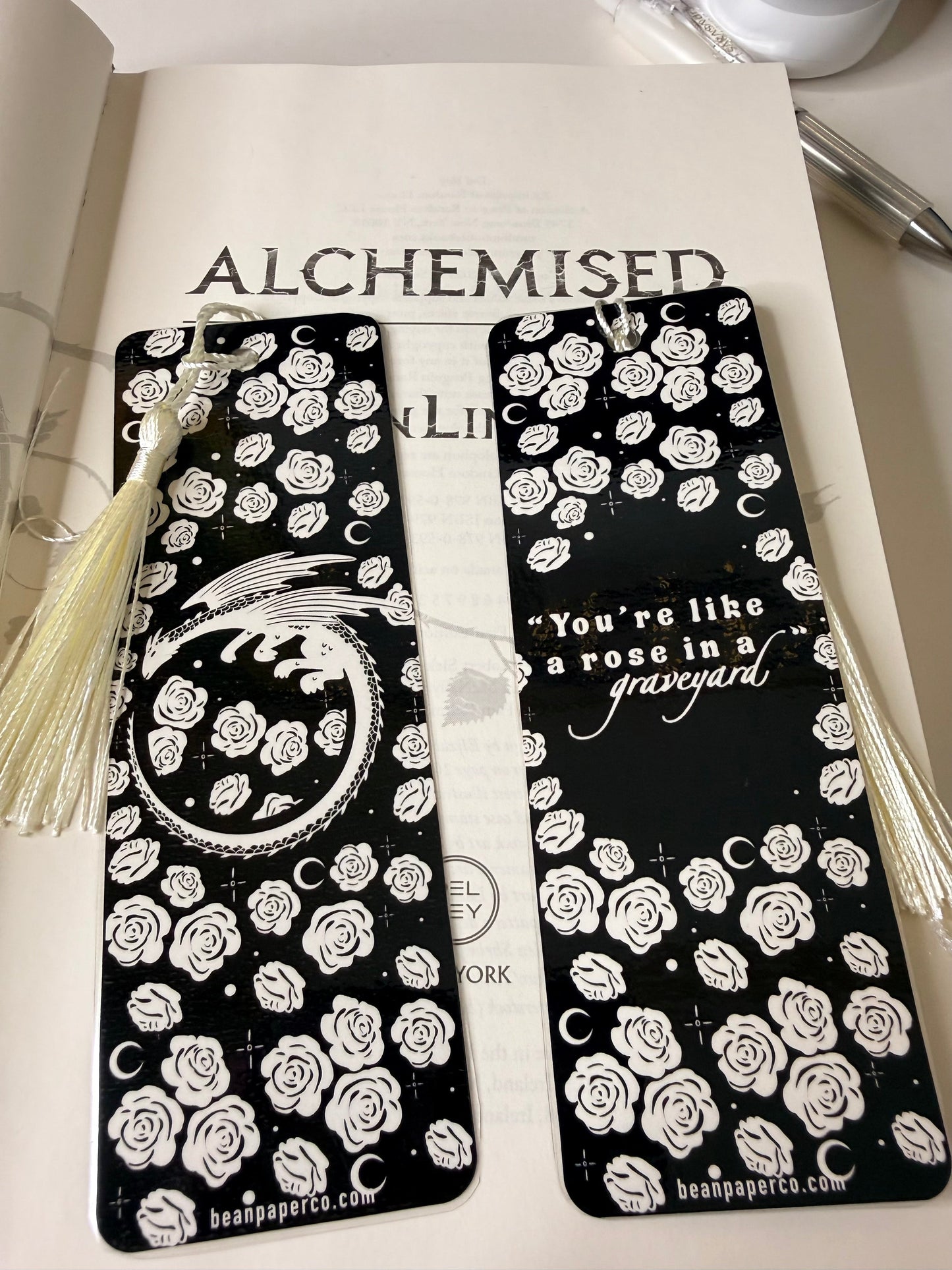 Two black bookmarks with white rose pattern and cream tassels resting on an open book, one featuring a dragon illustration and the other a romantic quote.