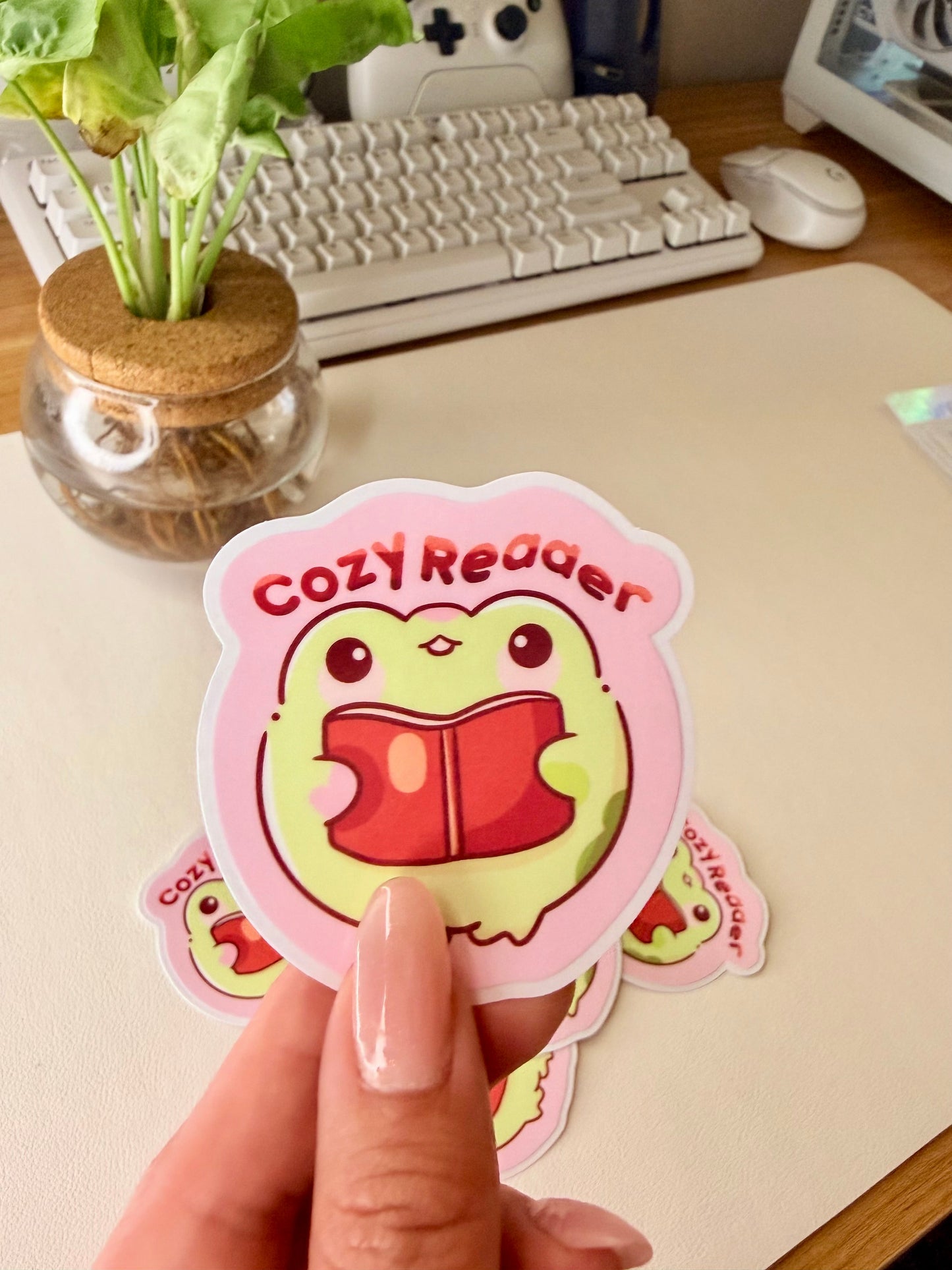 “Cute frog ‘Cozy Reader’ sticker featuring a green frog holding a red book on a pink background.”