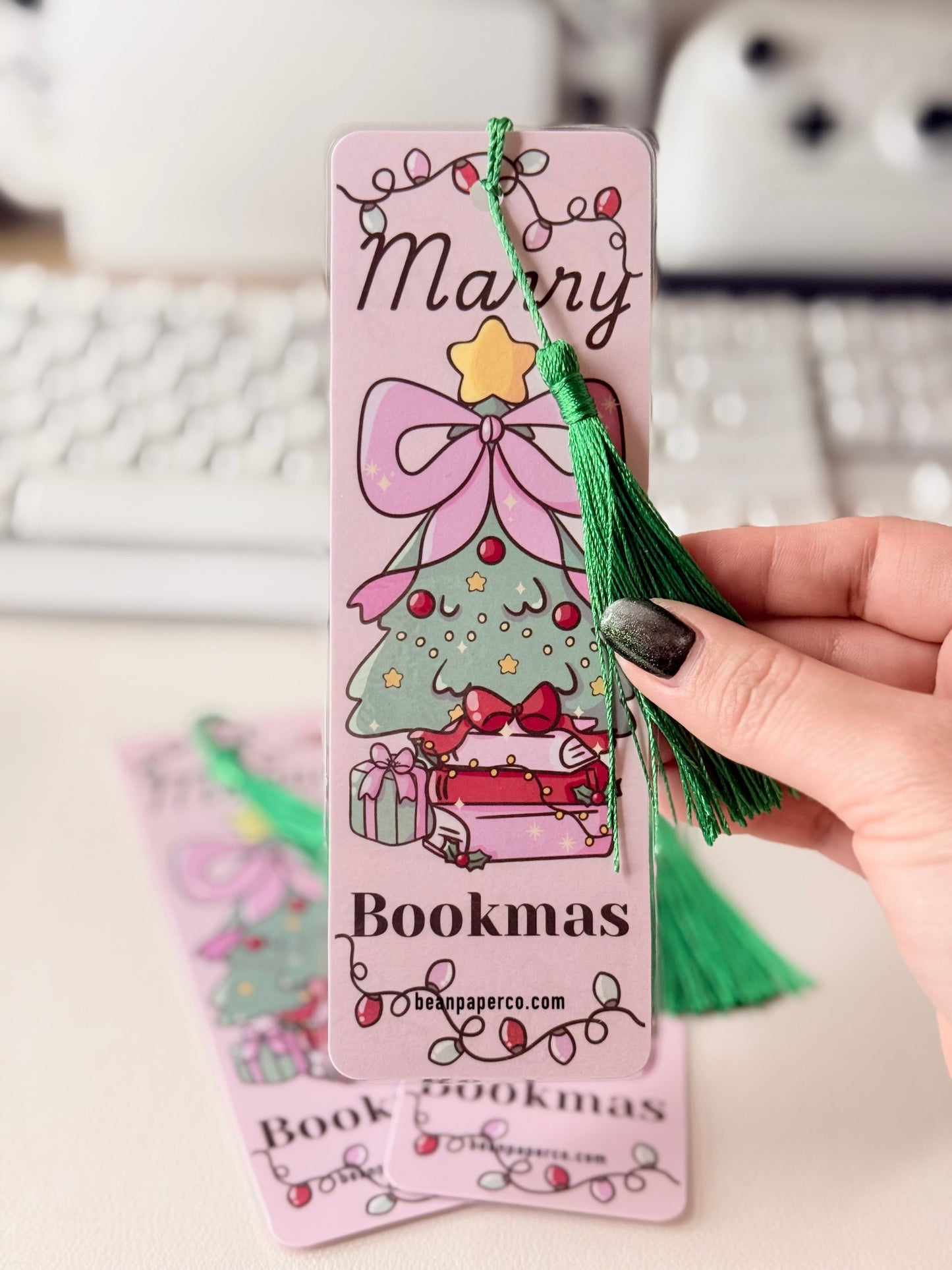 “Close-up of pink Merry Bookmas bookmark with green tassel and illustrated Christmas tree on stacked books.”