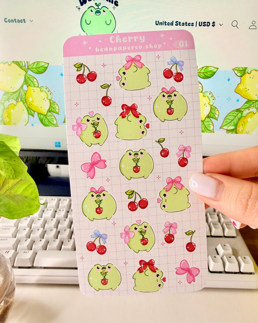 Hand holding a Cherry-themed frog sticker sheet featuring cute green frogs with pink bows, and red cherries.