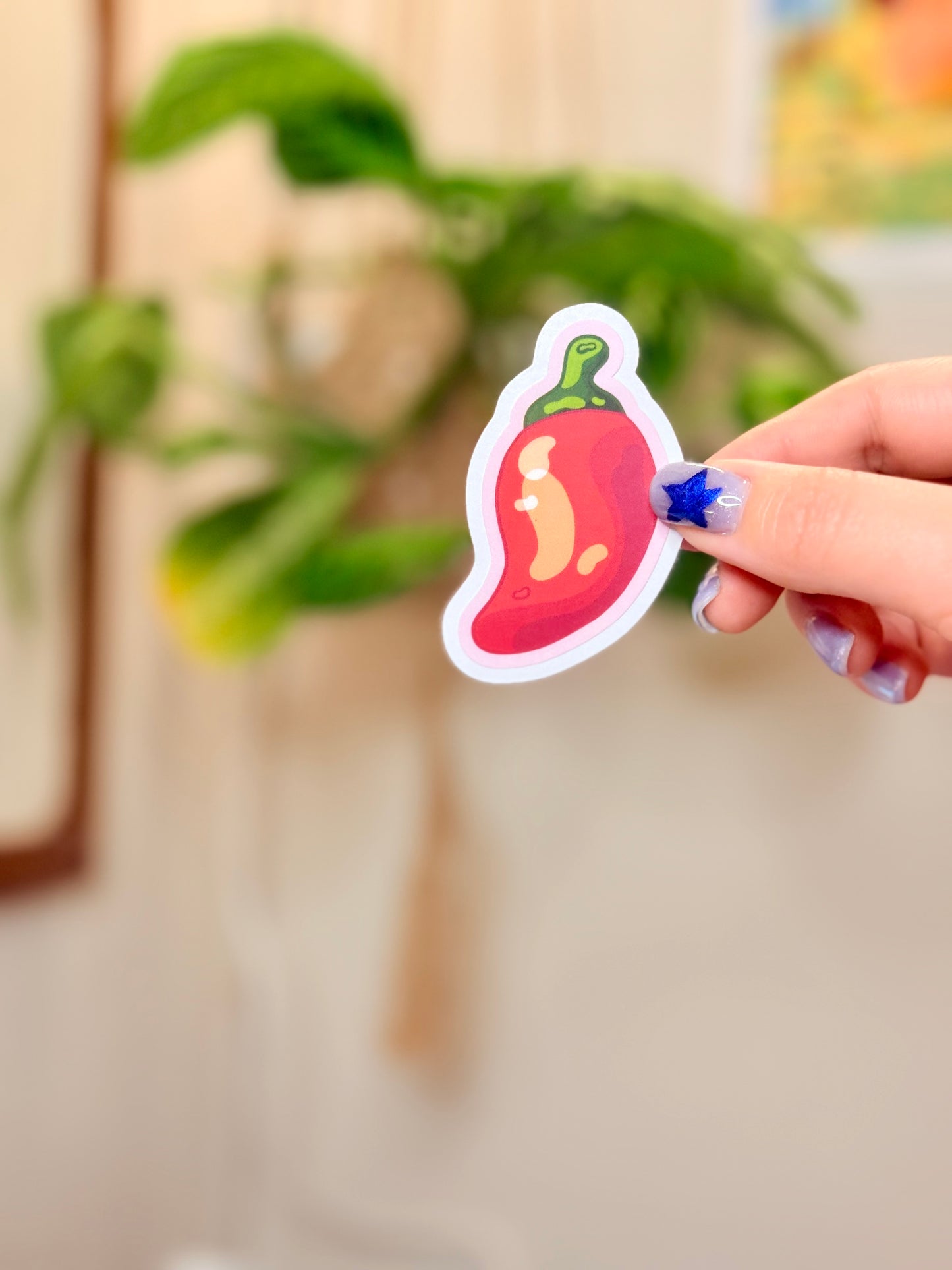 Hand holding a sticker of a chili pepper with pink outline against a blurred indoor background