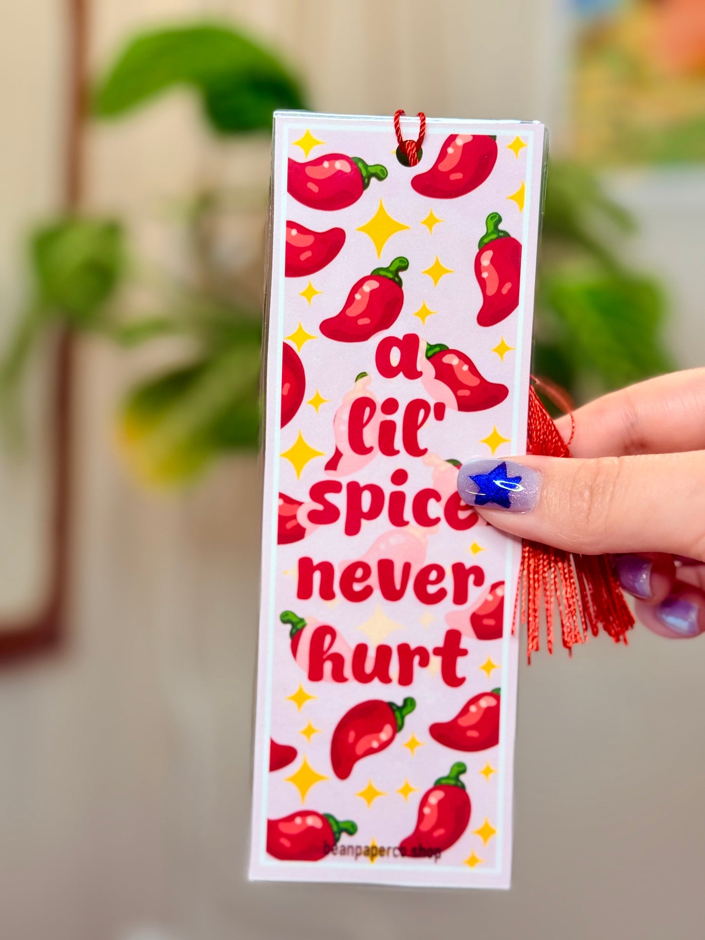 Bookmark with red peppers, a red tassel and text that says "a lil spice never hurt," held by a hand, with a plant in the background