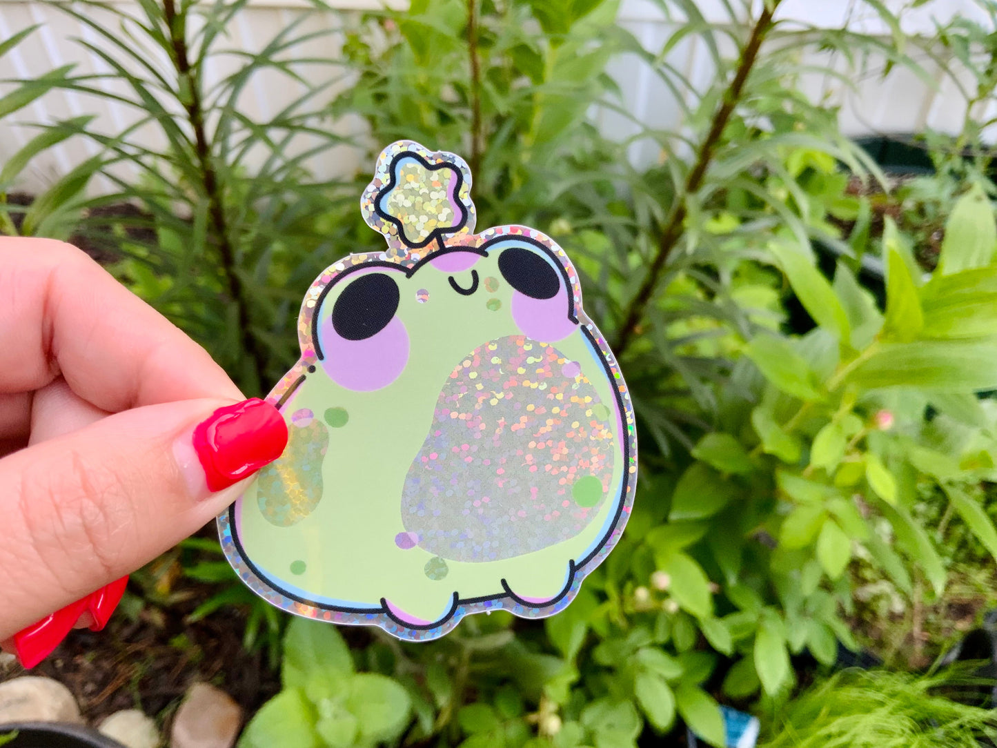 Hand holding a glittery green frog sticker with purple cheeks and a sparkly star detail, photographed outdoors against leafy green plants.