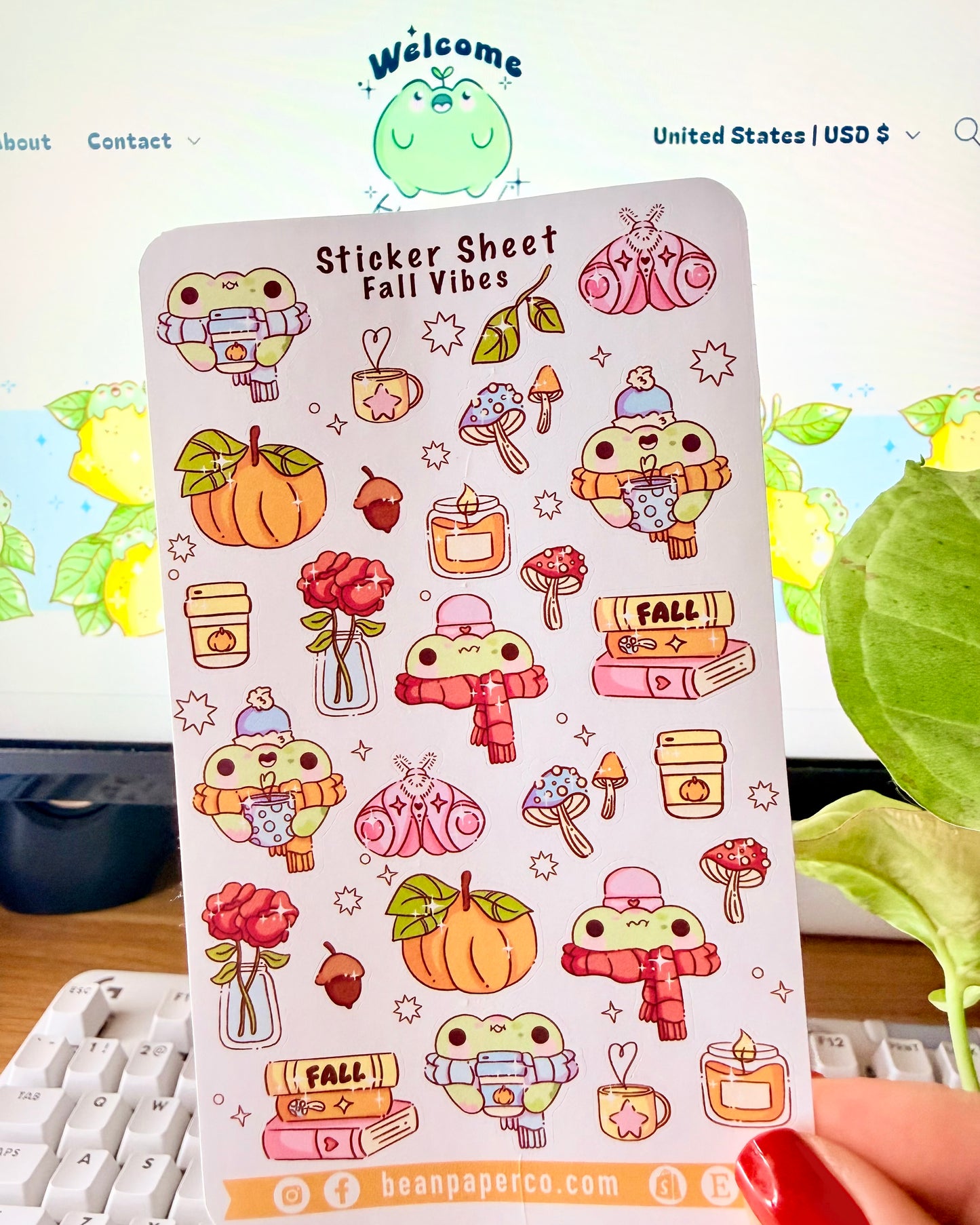Sticker sheet titled “Sticker Sheet Fall Vibes” featuring cute frog illustrations with pumpkins, scarves, mushrooms, candles, books, acorns, roses, and cozy autumn drinks, held in front of a desk setup.