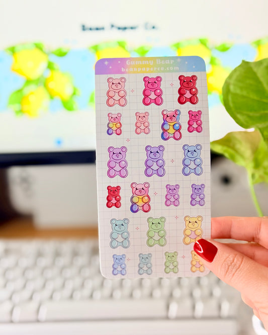 Valentine Gummy Bear Sticker Sheet. Hand holding a pastel gummy bear sticker sheet with multiple colorful bear designs in pink, purple, blue, green, and red on a grid background, photographed in front of a computer screen and plant.