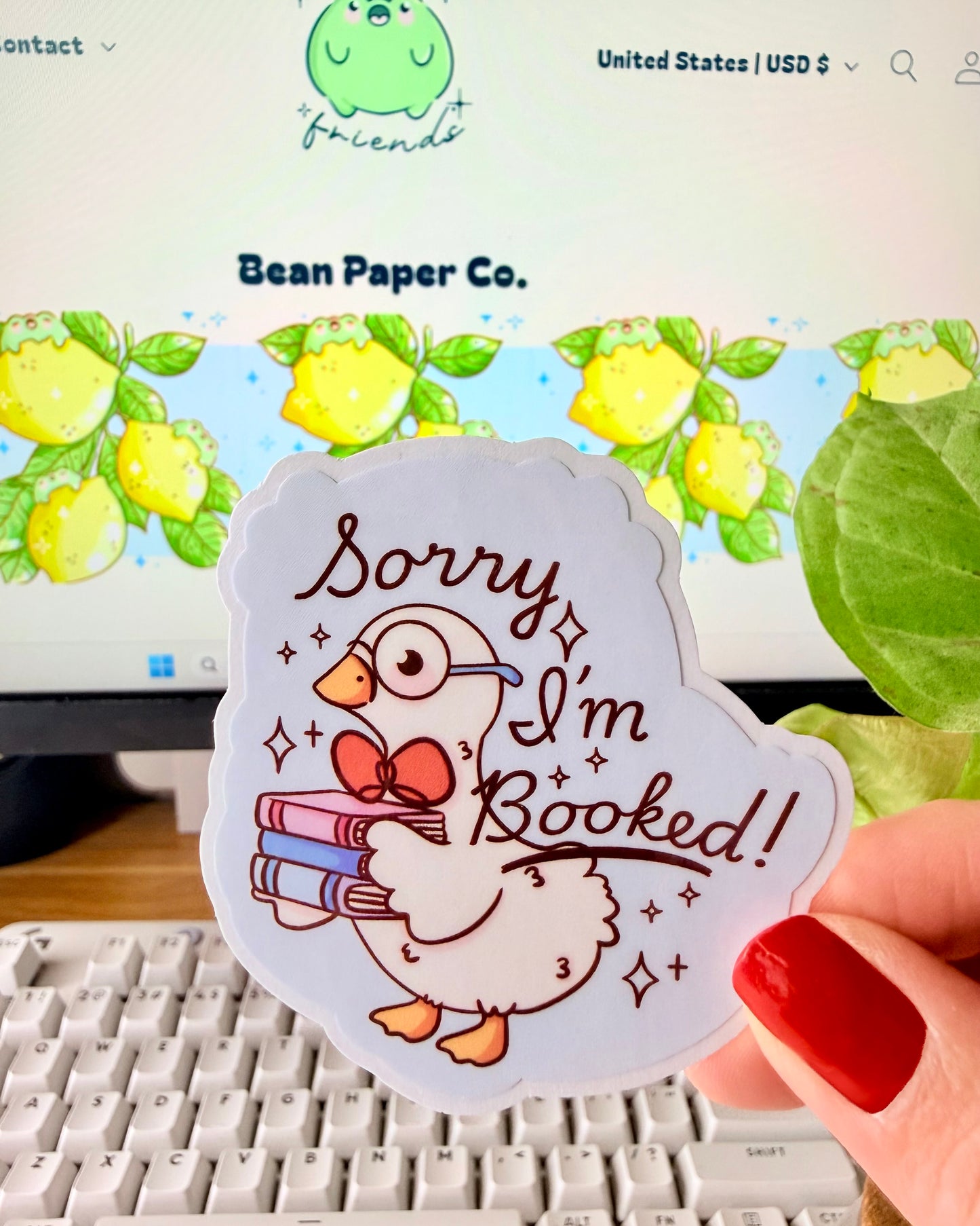 Adorable I'm Booked Duck Sticker. Hand holding a pastel vinyl sticker of a white goose wearing glasses and holding a stack of books with the text “Sorry I’m Booked!” in front of a computer keyboard and plant.