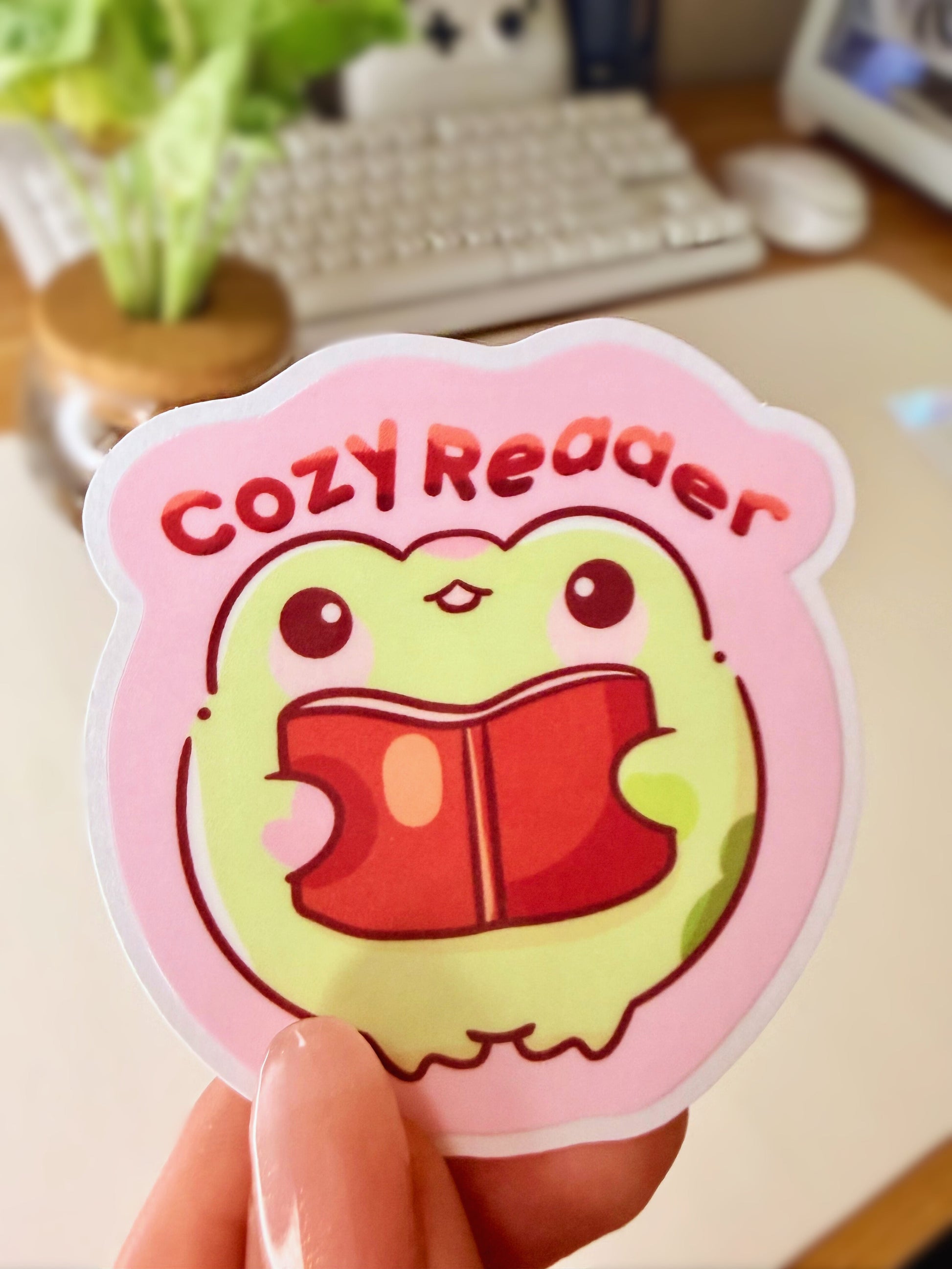 “Cute frog ‘Cozy Reader’ sticker featuring a green frog holding a red book on a pink background.”