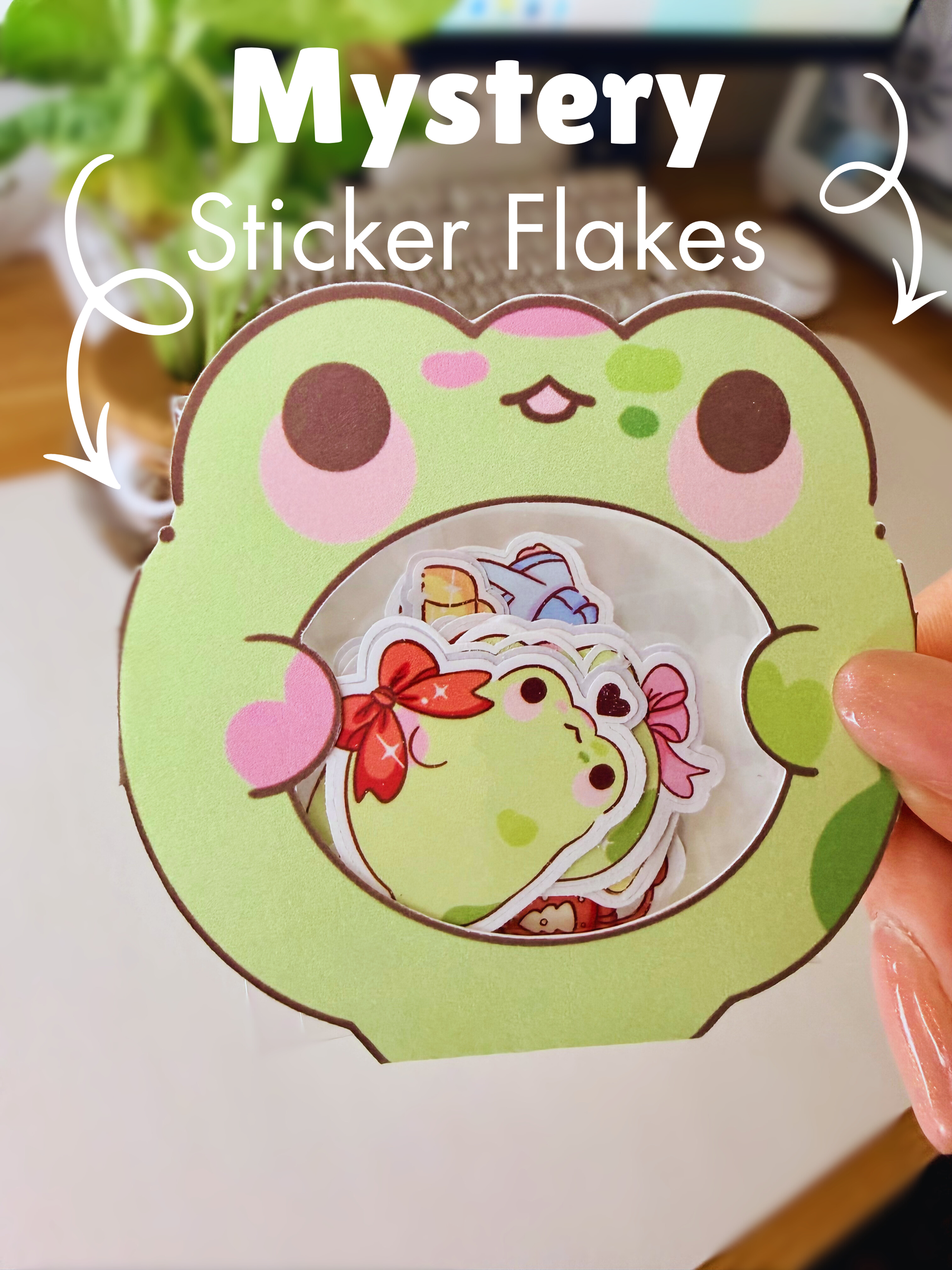 “Frog-shaped packaging holding small die-cut frog sticker flakes on a desk setup with the words Mystery Sticker Flakes and tow arrows on each side pointing to the frog."