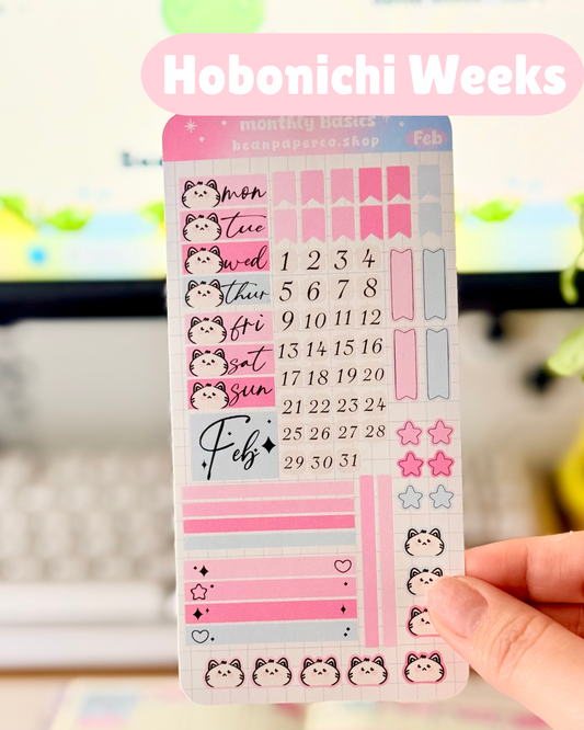 Hobonichi Weeks February Functional Monthly Planner Kitt featuring pink and blue date covers, weekday labels with cute cat faces, number stickers, star icons, and decorative boxes.