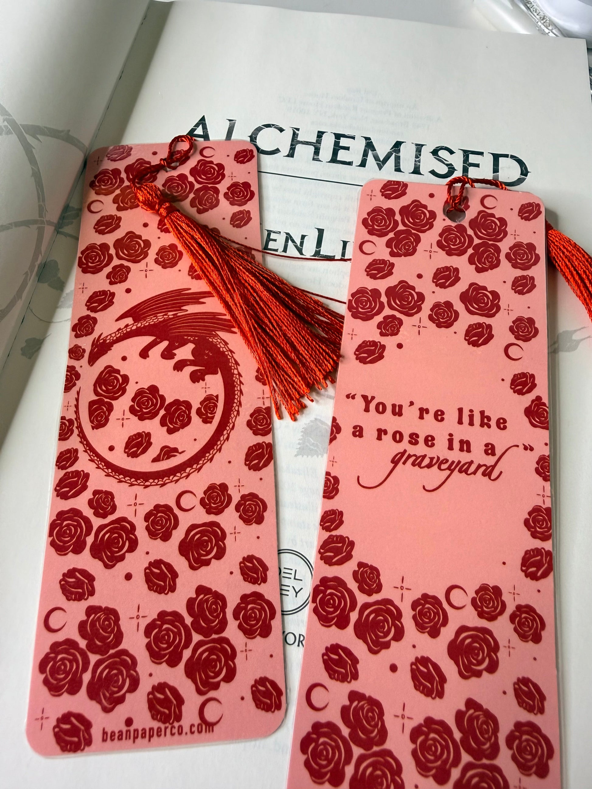 wo pink bookmarks with red rose pattern and red tassels resting on an open book, one featuring a dragon design and the other a romantic quote.
