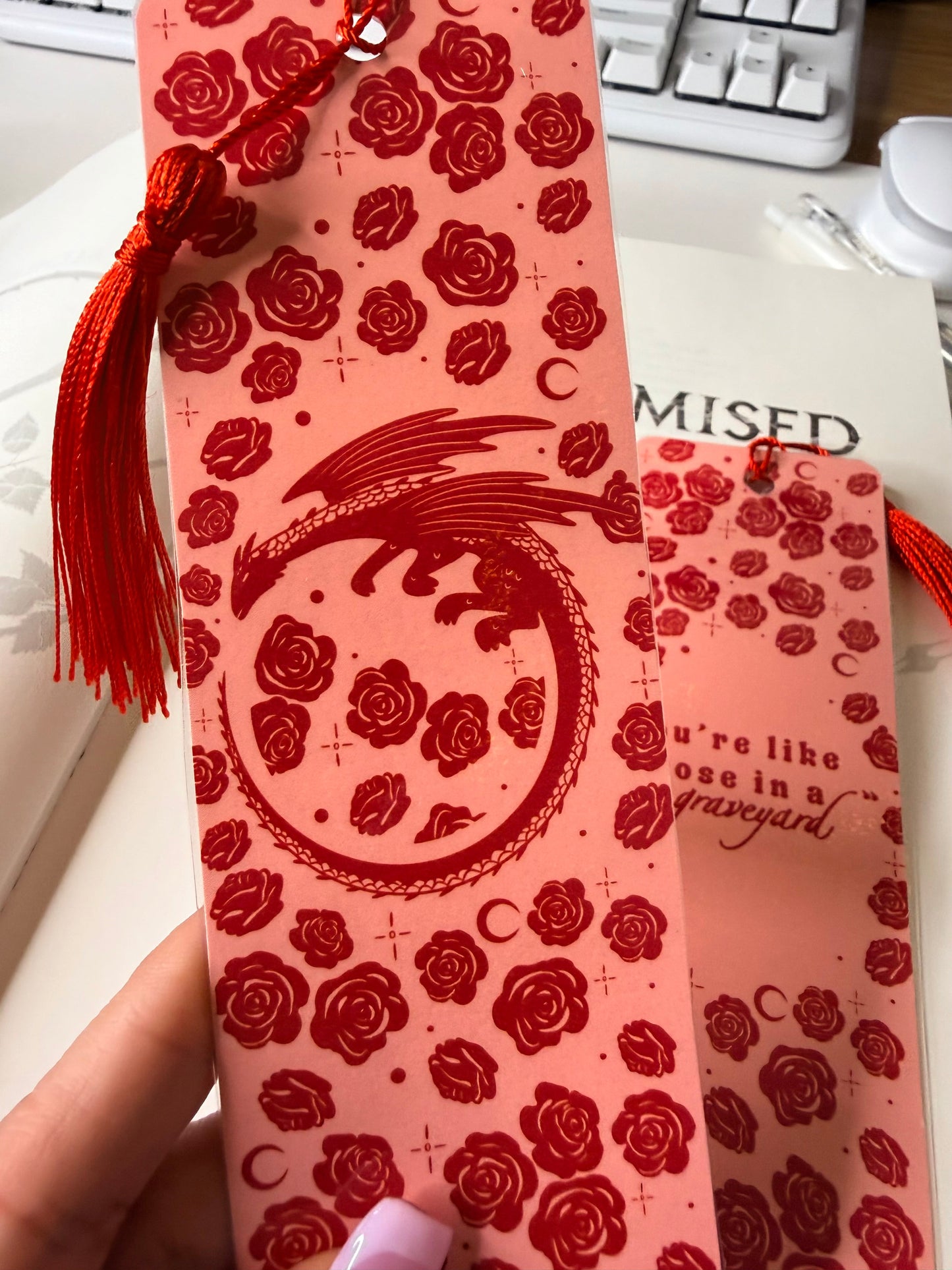 Pink bookmark with red rose pattern and circular dragon design, finished with a red tassel, held in front of an open book.