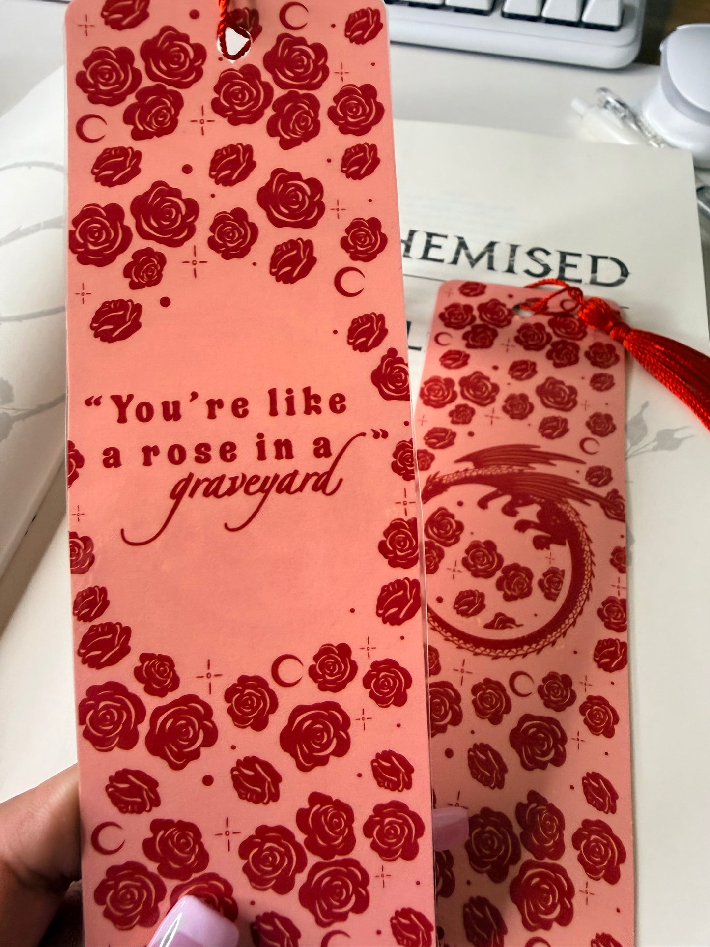 Pink bookmark with red roses and the quote “You’re like a rose in a graveyard” in red lettering, shown against an open book.