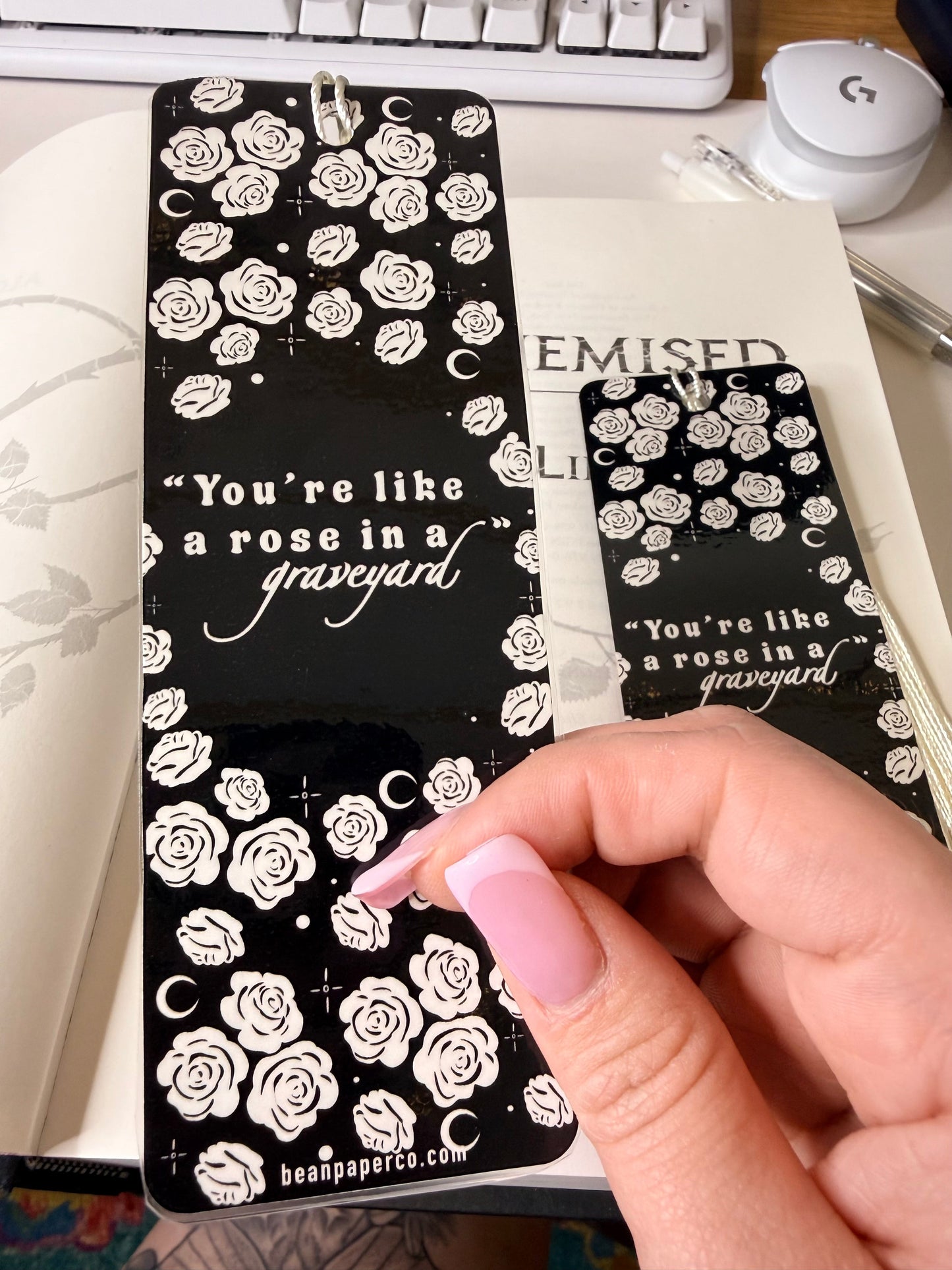 Black bookmark with white roses and the quote “You’re like a rose in a graveyard” in white lettering, shown against an open book.