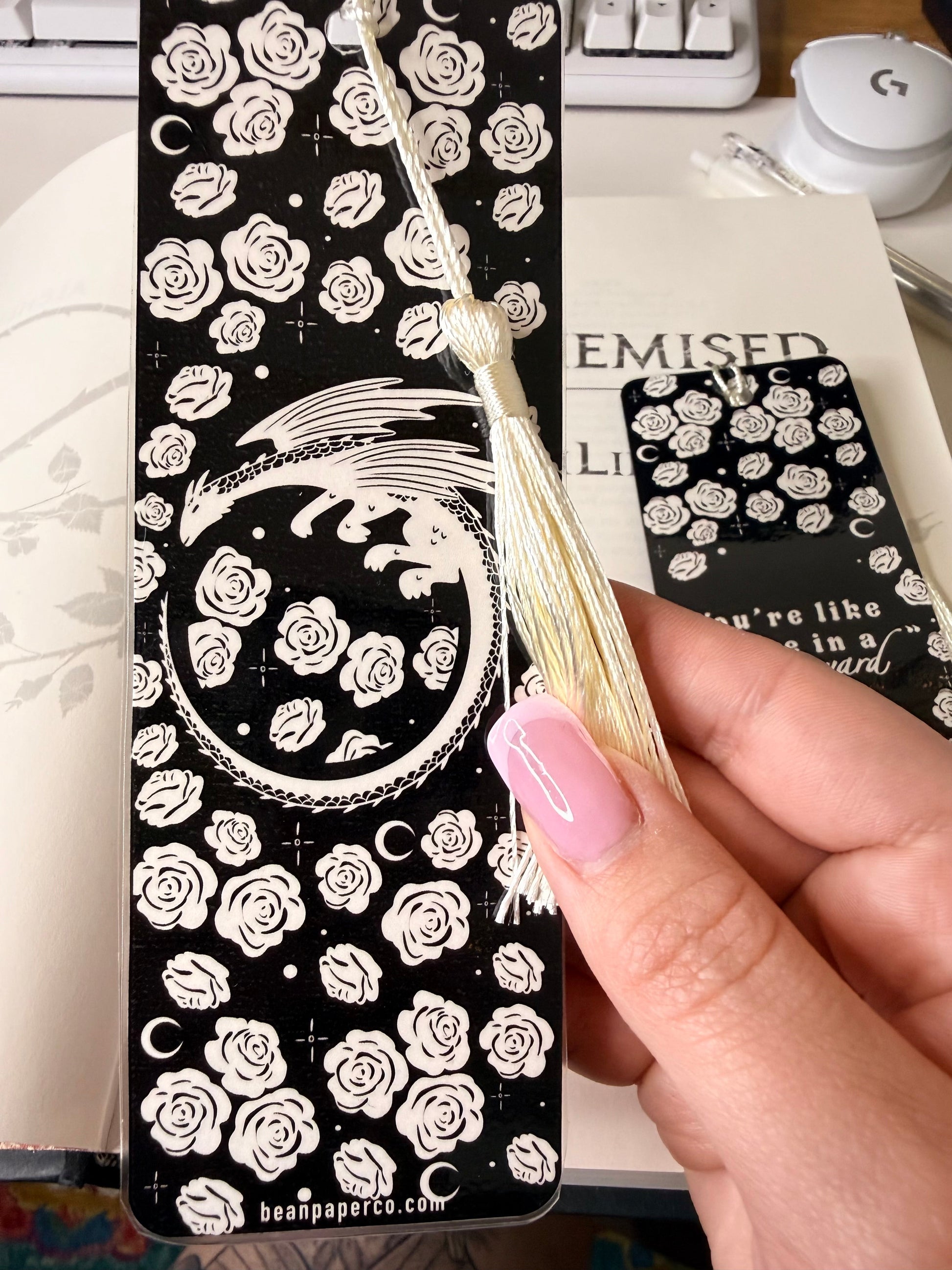 Black bookmark with white rose pattern and circular dragon design, finished with a cream tassel, held over an open book.