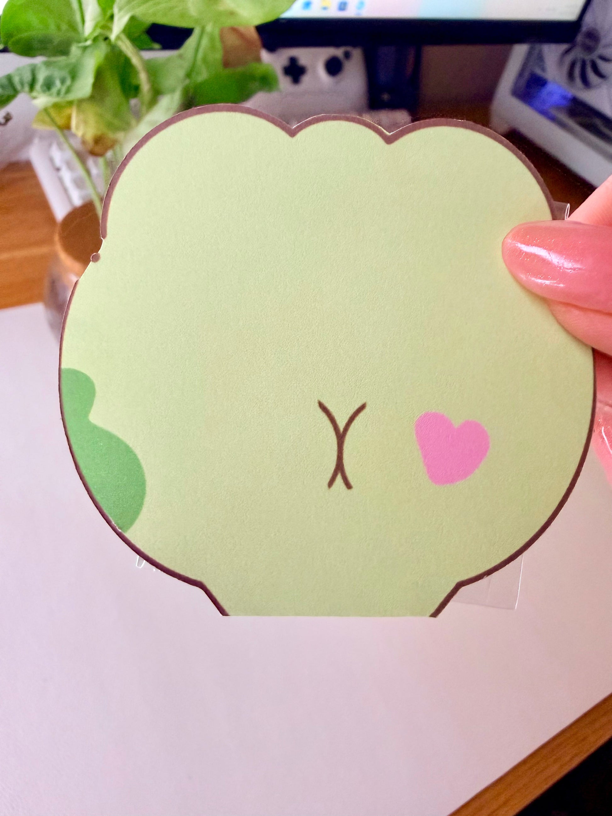 “Frog-shaped sticker flake pouch shown from the back, featuring a pastel green frog design with a pink heart detail.”
