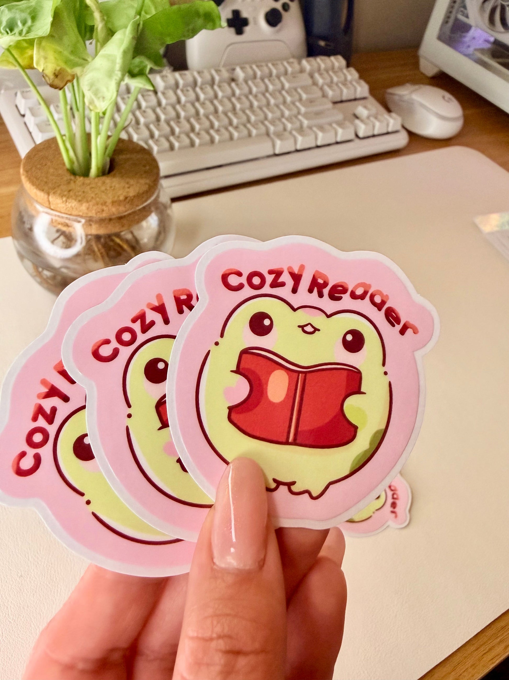 “Cute frog ‘Cozy Reader’ sticker featuring a green frog holding a red book on a pink background.”
