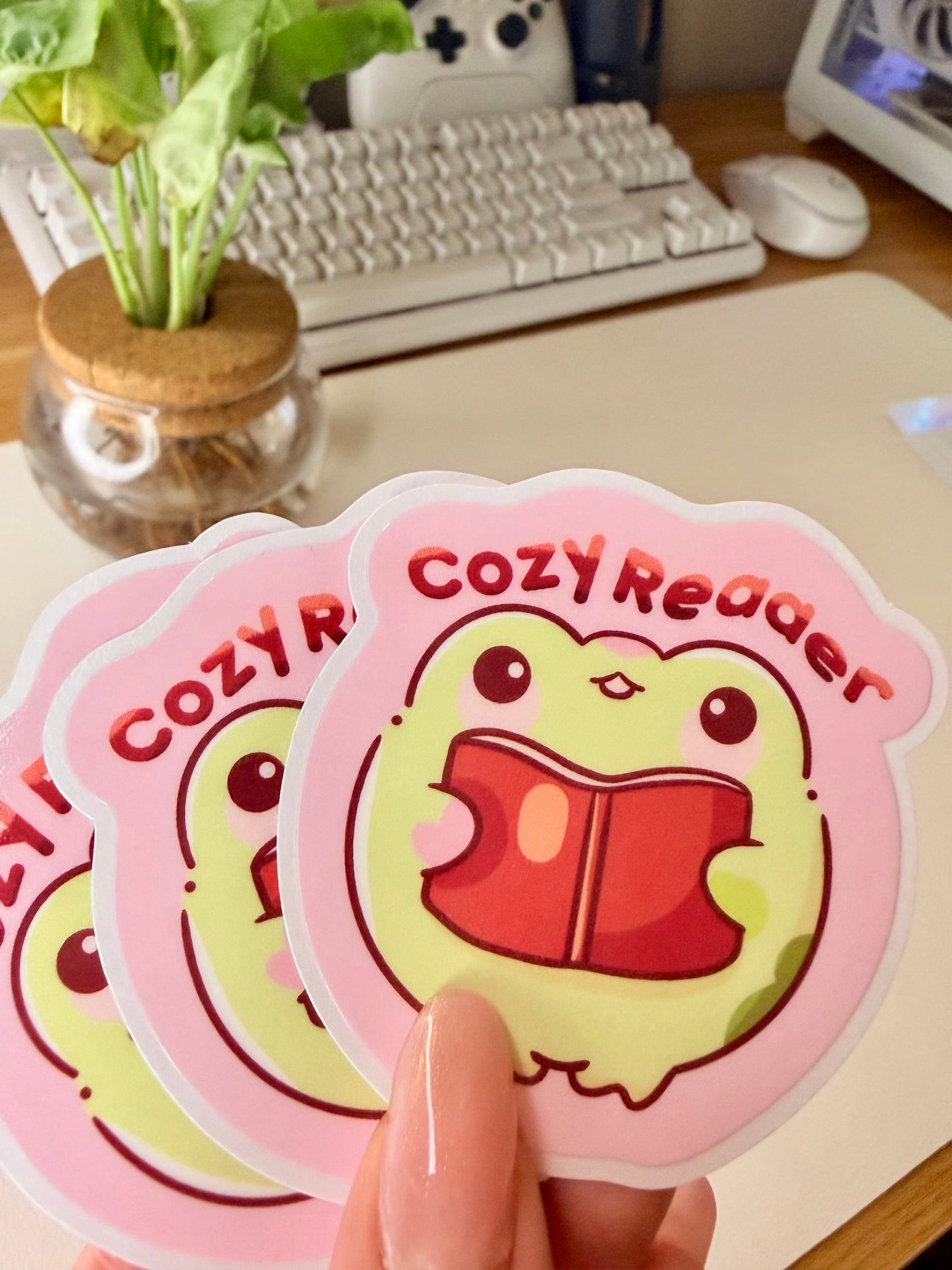 “Cute frog ‘Cozy Reader’ sticker featuring a green frog holding a red book on a pink background.”