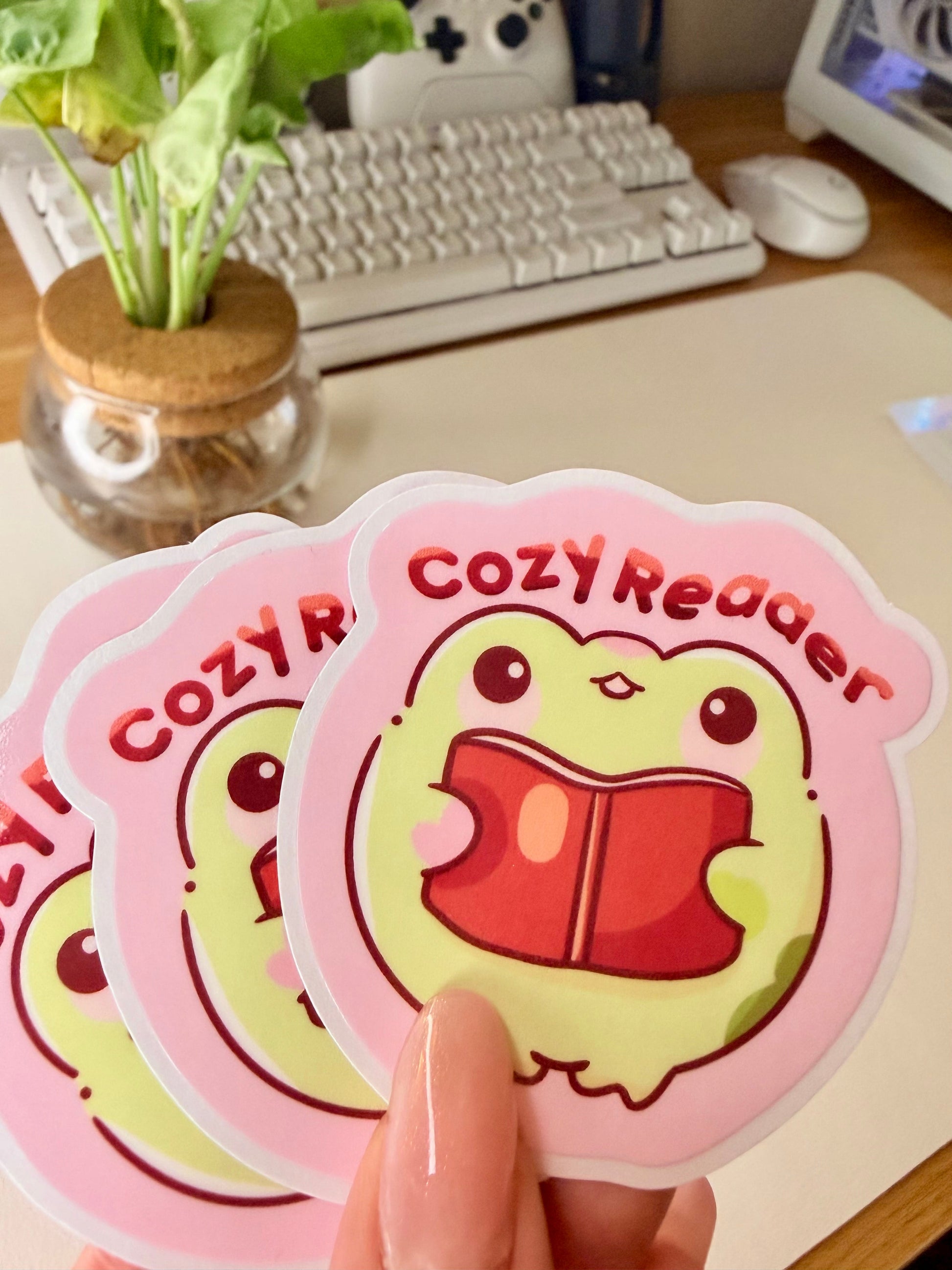 “Cute frog ‘Cozy Reader’ sticker featuring a green frog holding a red book on a pink background.”