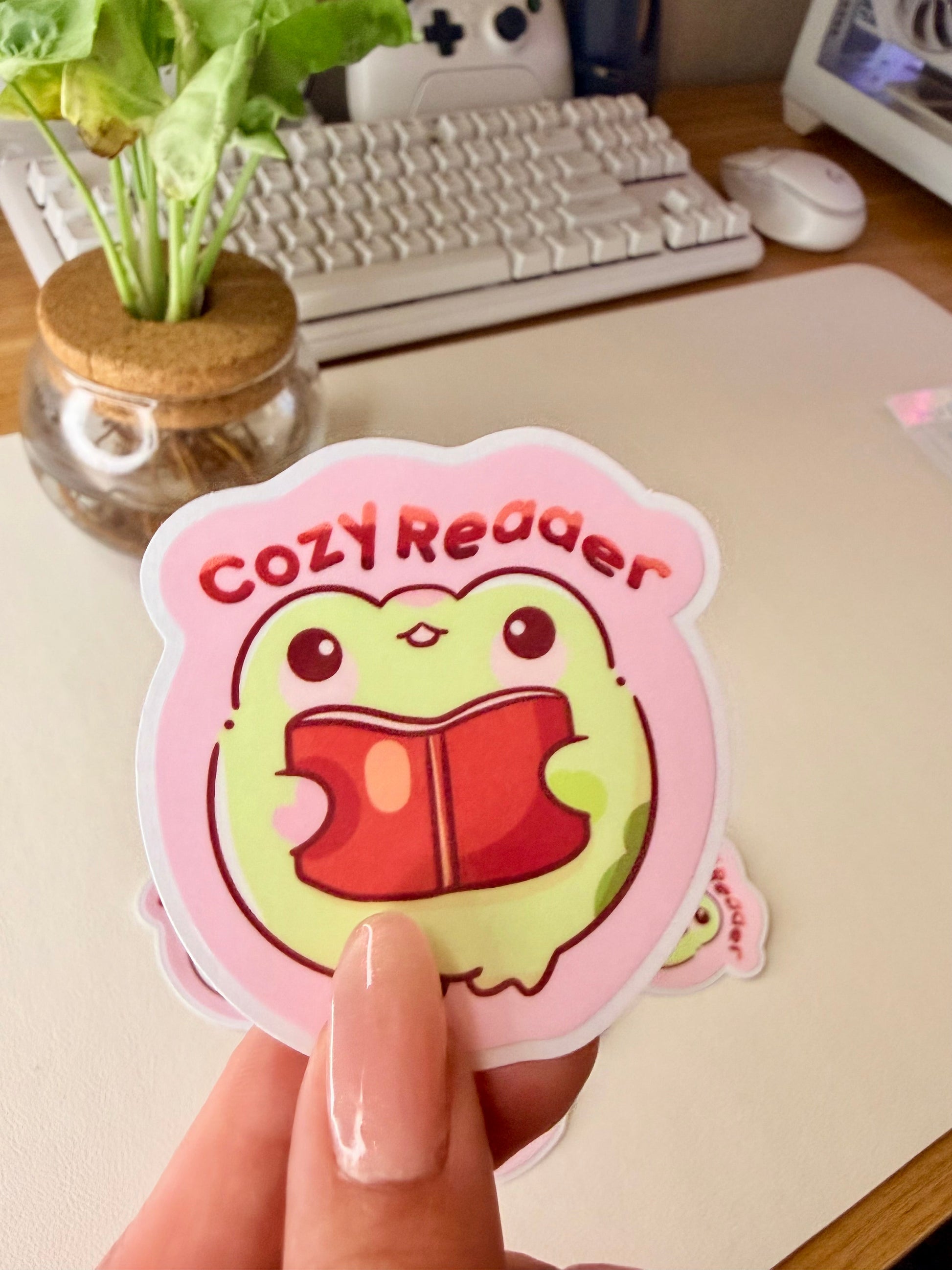 “Cute frog ‘Cozy Reader’ sticker featuring a green frog holding a red book on a pink background.”