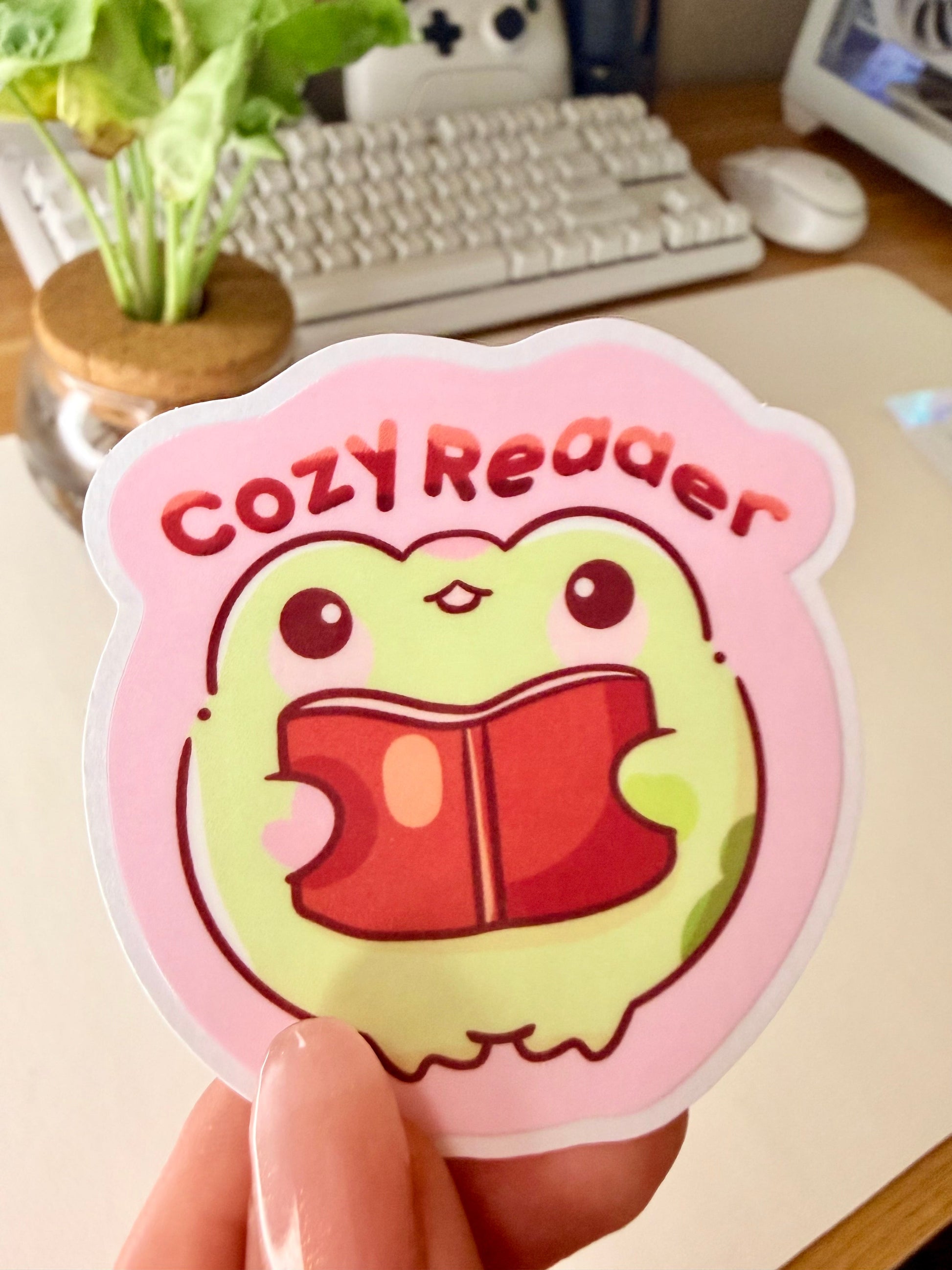“Cute frog ‘Cozy Reader’ sticker featuring a green frog holding a red book on a pink background.”