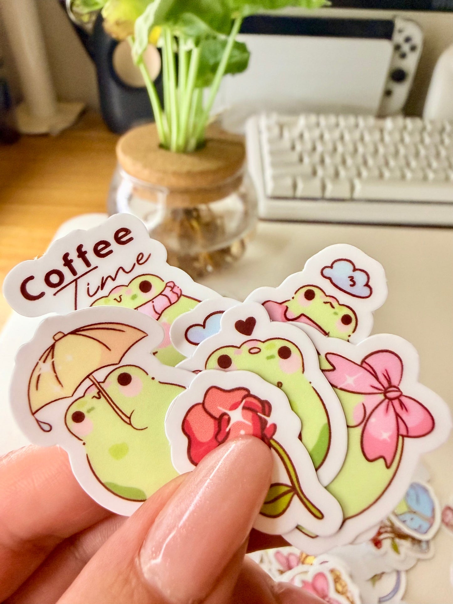 “Hand holding assorted frog sticker flakes featuring bows, flowers, cherries, and cozy-themed frog designs.”