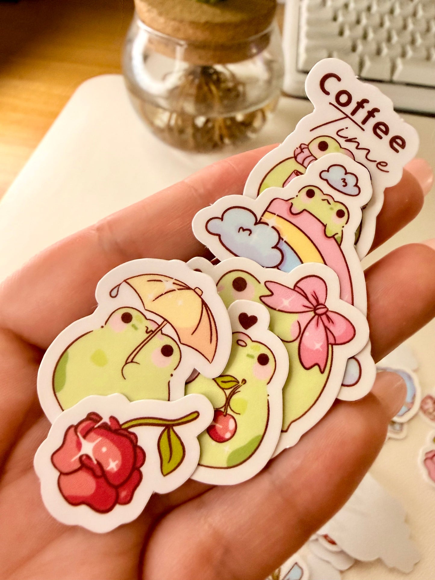 “Hand holding assorted frog sticker flakes featuring bows, flowers, cherries, and cozy-themed frog designs.”