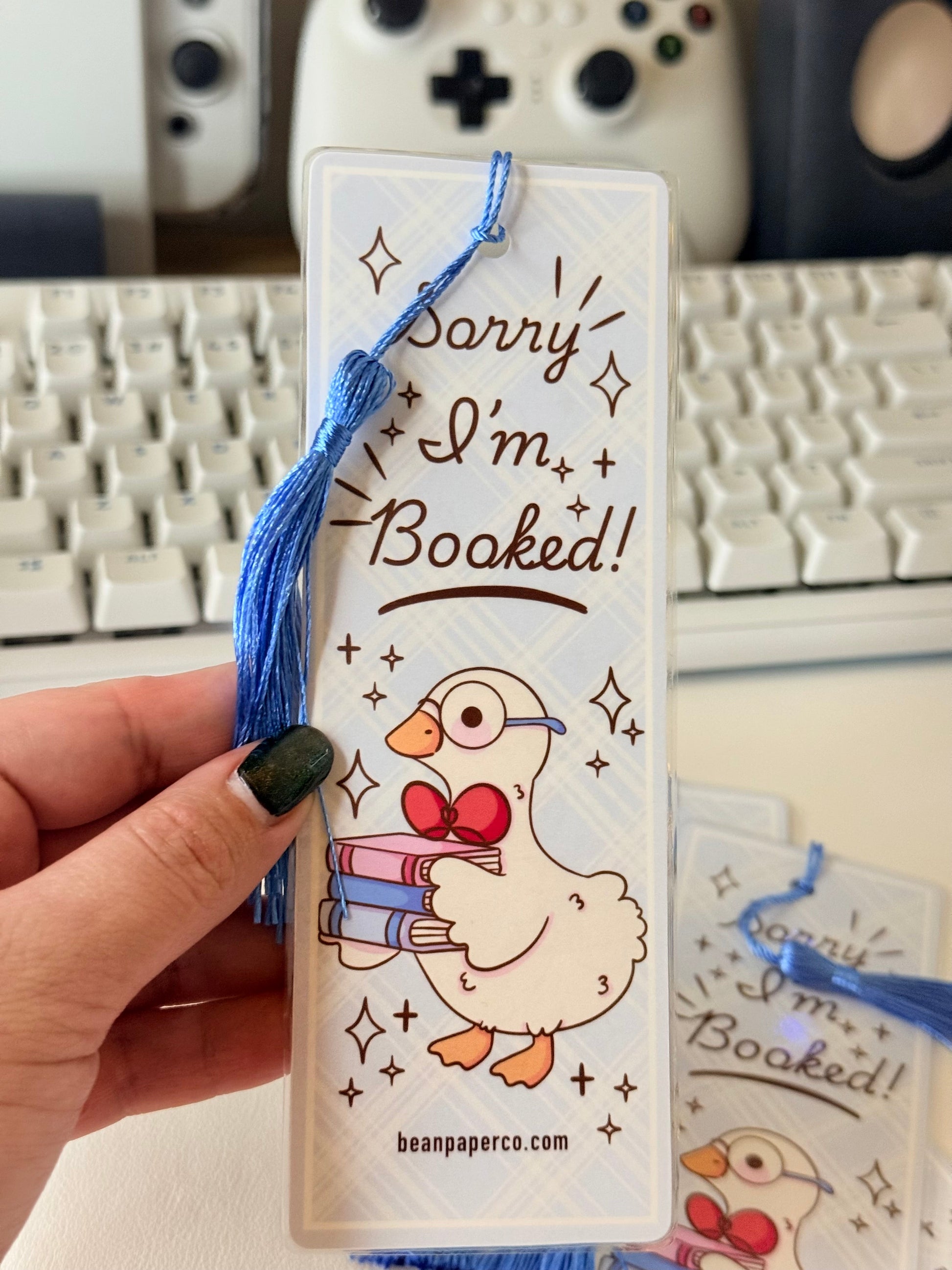 “Laminated bookmark with blue tassel featuring a duck holding books and the phrase ‘Sorry I’m Booked.’”