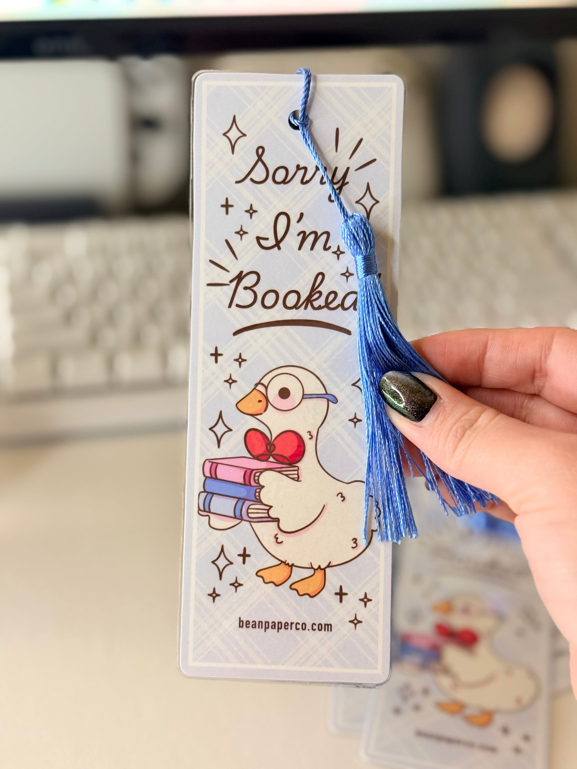 “Laminated bookmark with blue tassel featuring a duck holding books and the phrase ‘Sorry I’m Booked.’”