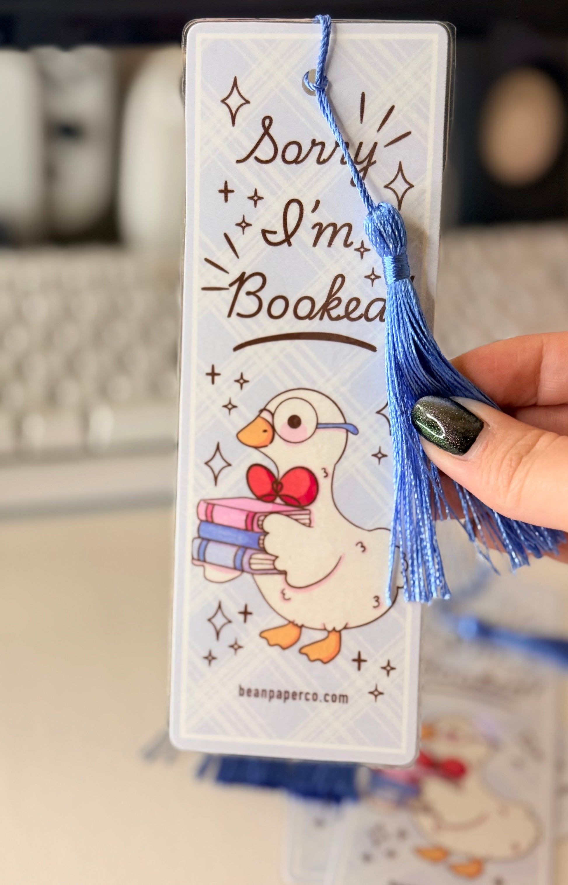 “Laminated bookmark with blue tassel featuring a duck holding books and the phrase ‘Sorry I’m Booked.’”