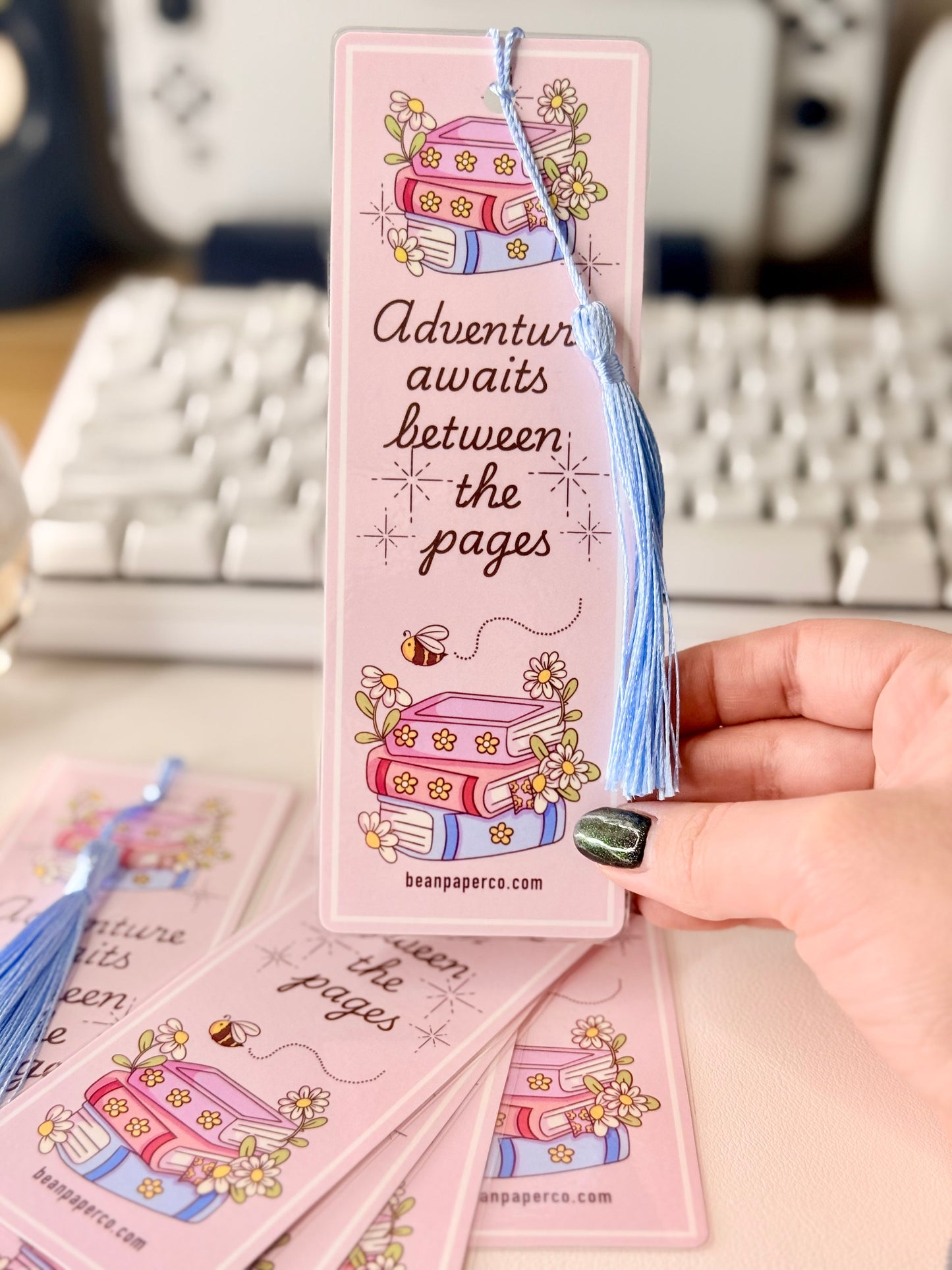 “Pink laminated bookmark with blue tassel featuring stacked books, daisies, and the phrase ‘Adventure awaits between the pages.’”