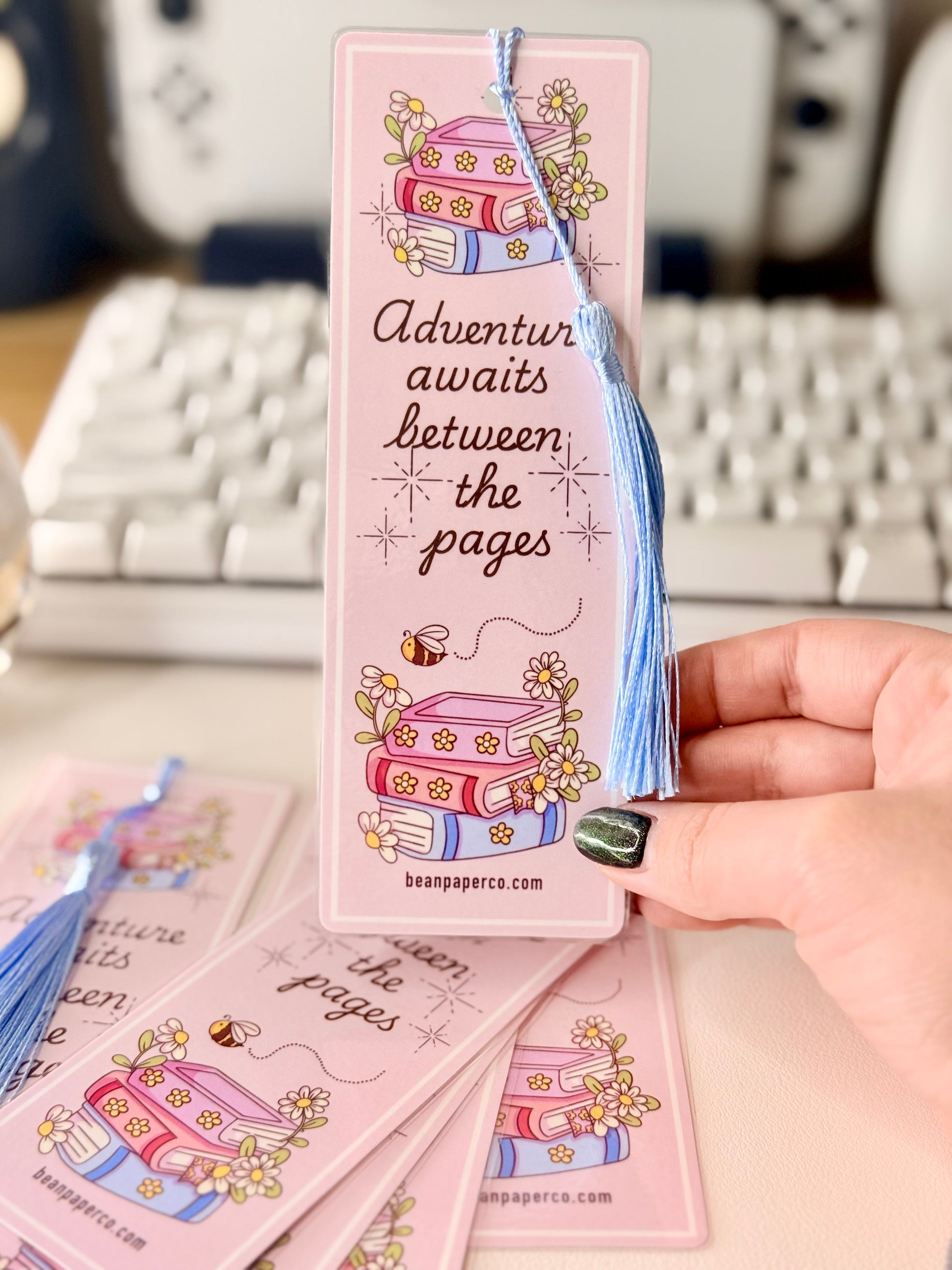 “Pink laminated bookmark with blue tassel featuring stacked books, daisies, and the phrase ‘Adventure awaits between the pages.’”