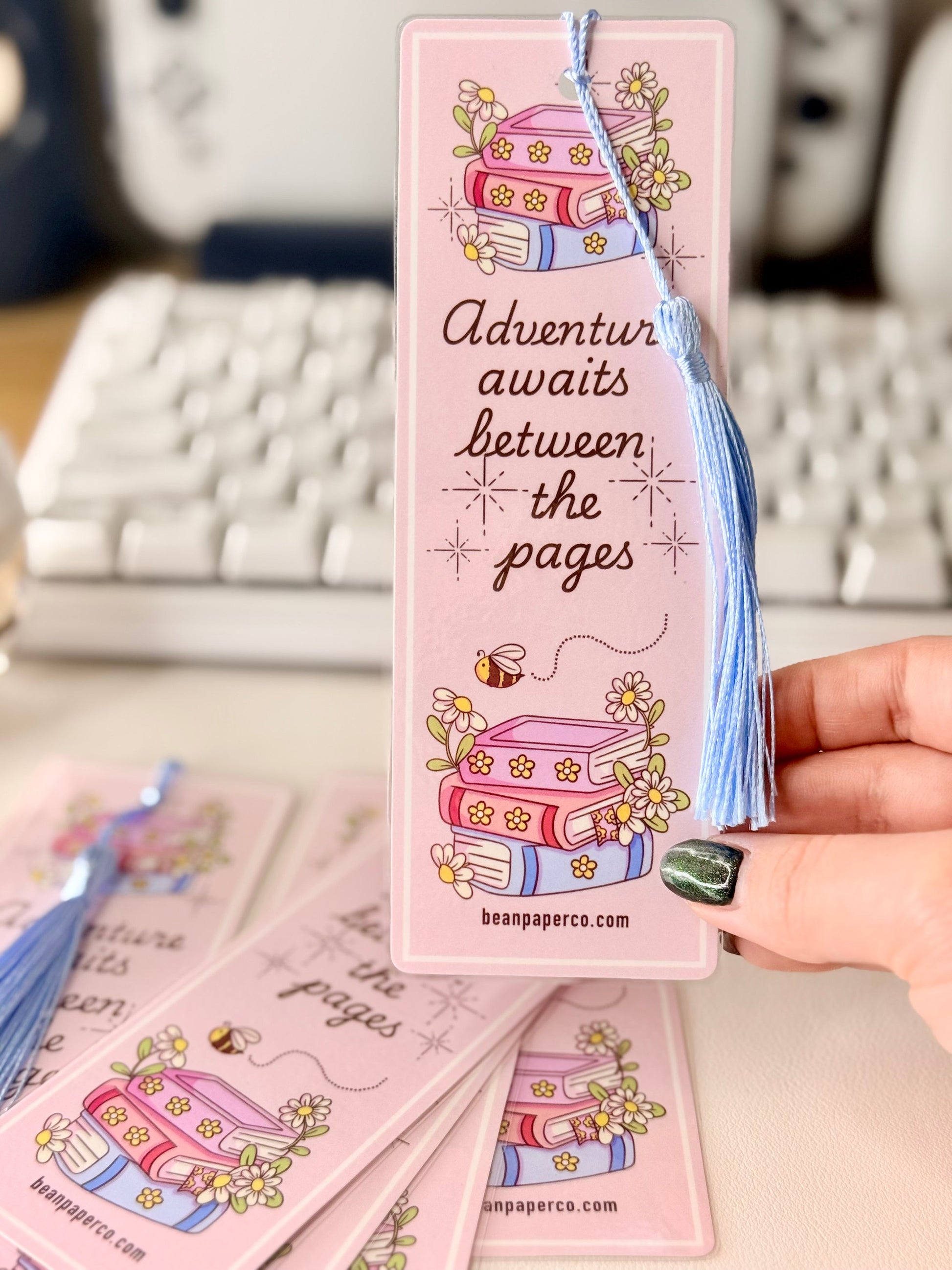 “Pink laminated bookmark with blue tassel featuring stacked books, daisies, and the phrase ‘Adventure awaits between the pages.’”