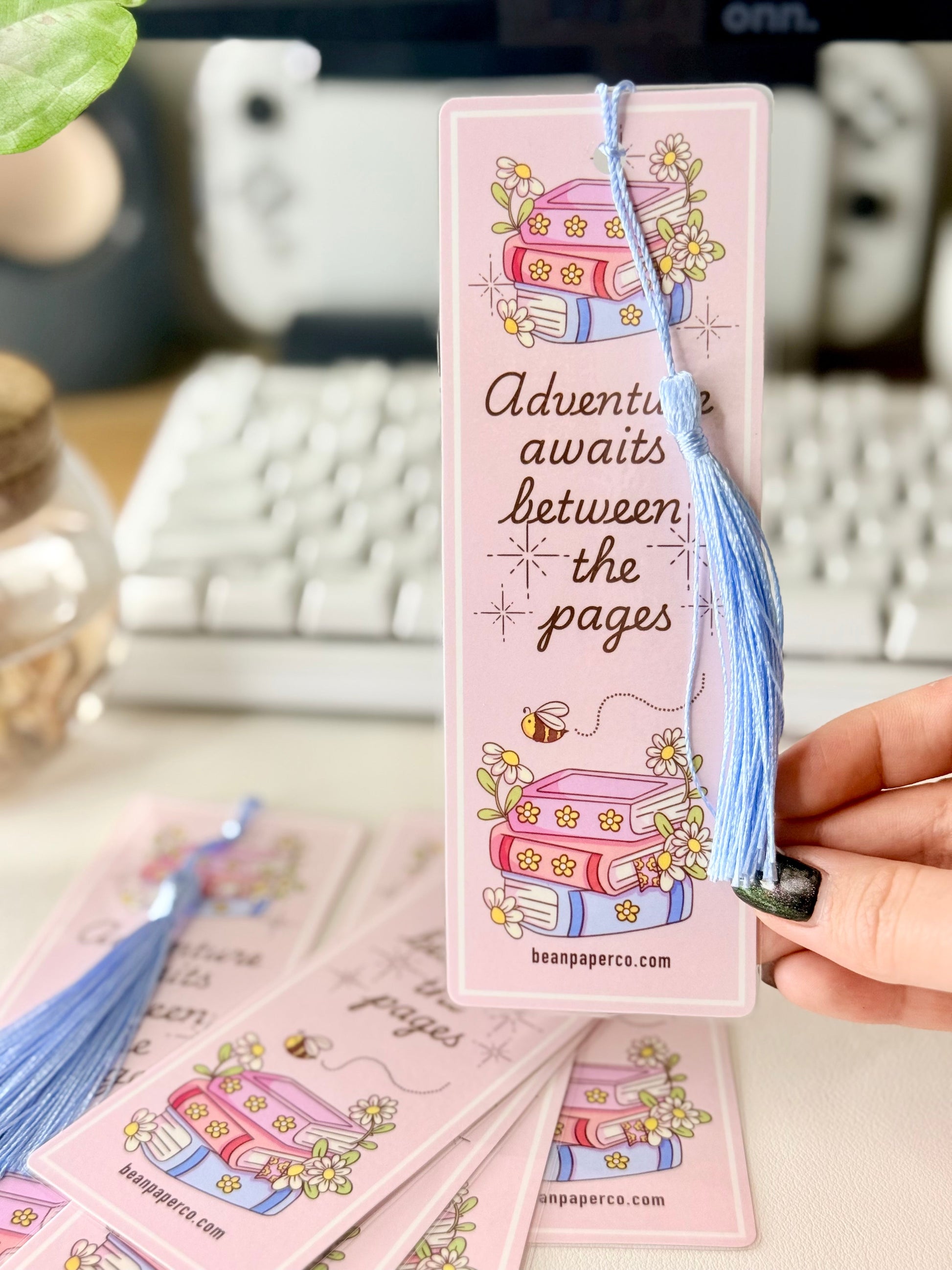 “Pink laminated bookmark with blue tassel featuring stacked books, daisies, and the phrase ‘Adventure awaits between the pages.’”