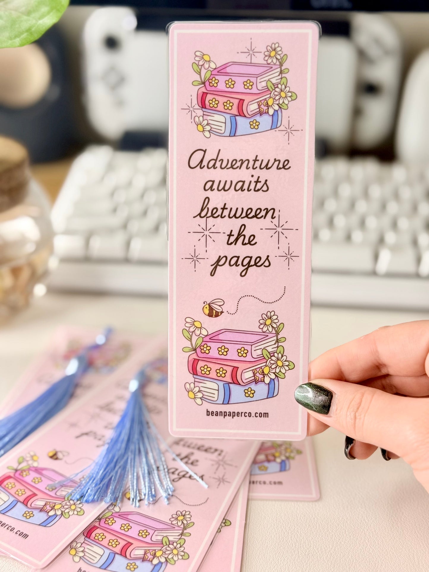 “Laminated pink bookmark featuring illustrated books with flowers and the quote ‘Adventure awaits between the pages.’”