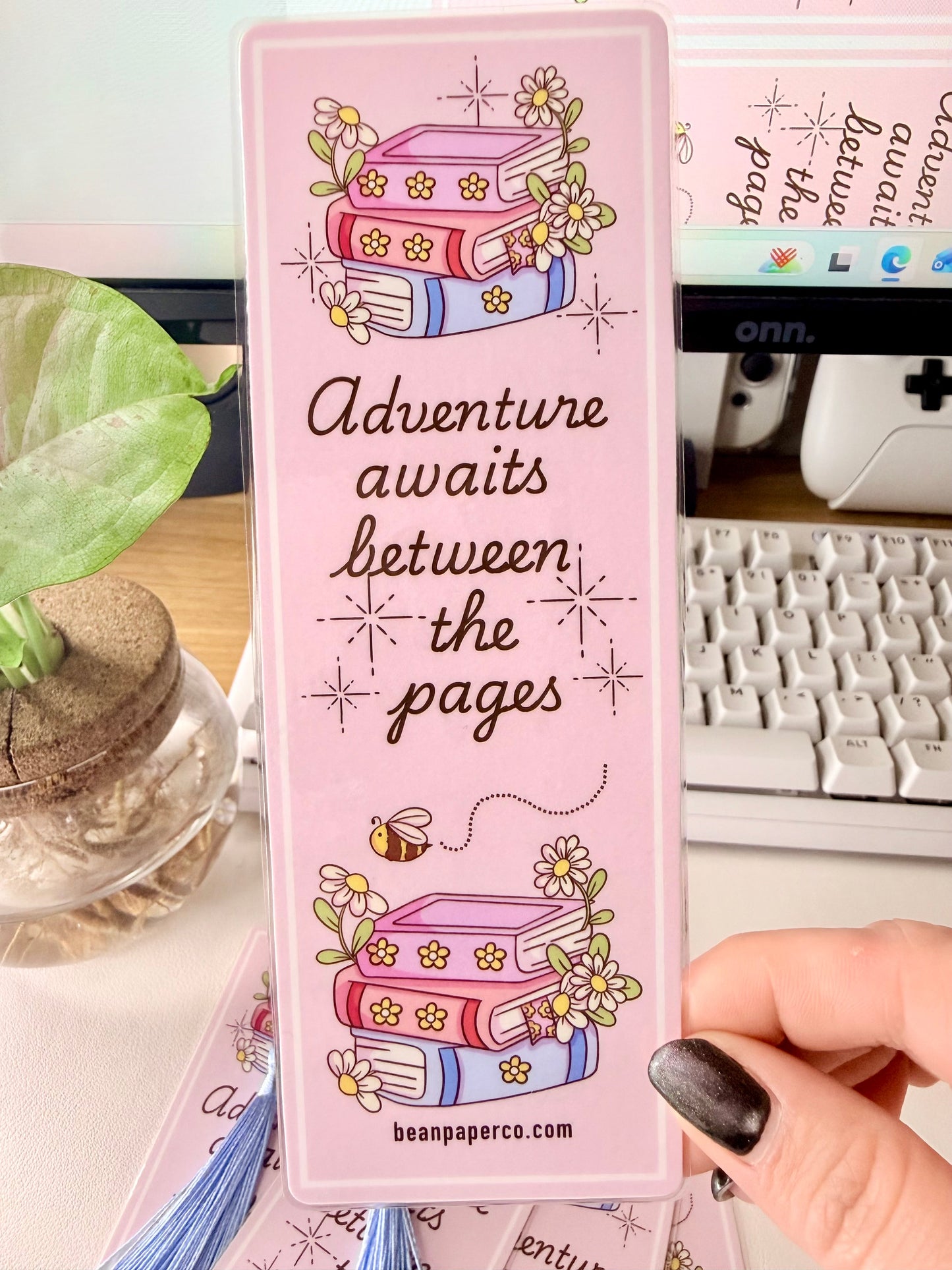 “Laminated pink bookmark featuring illustrated books with flowers and the quote ‘Adventure awaits between the pages.’”