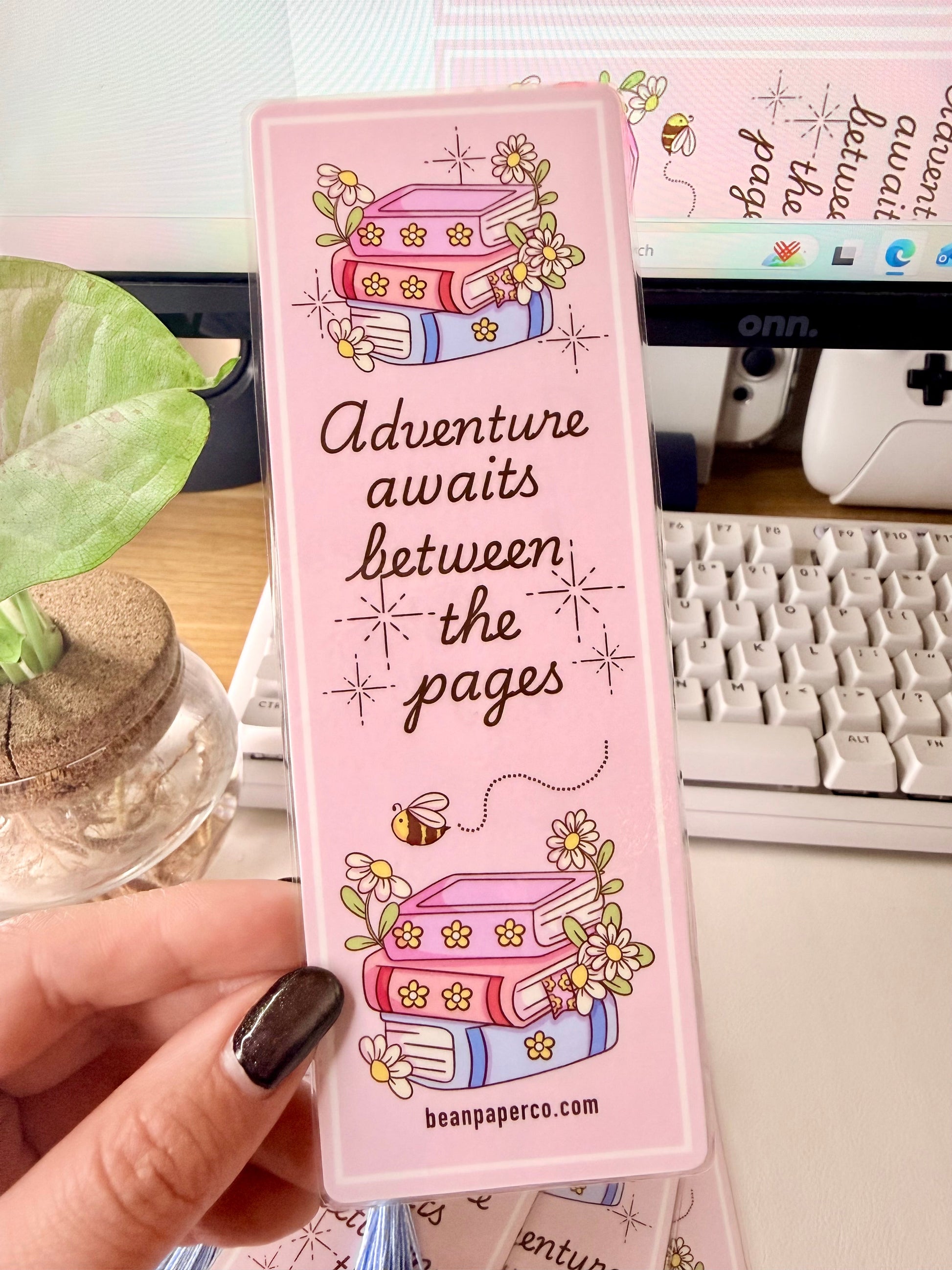“Laminated pink bookmark featuring illustrated books with flowers and the quote ‘Adventure awaits between the pages.’”