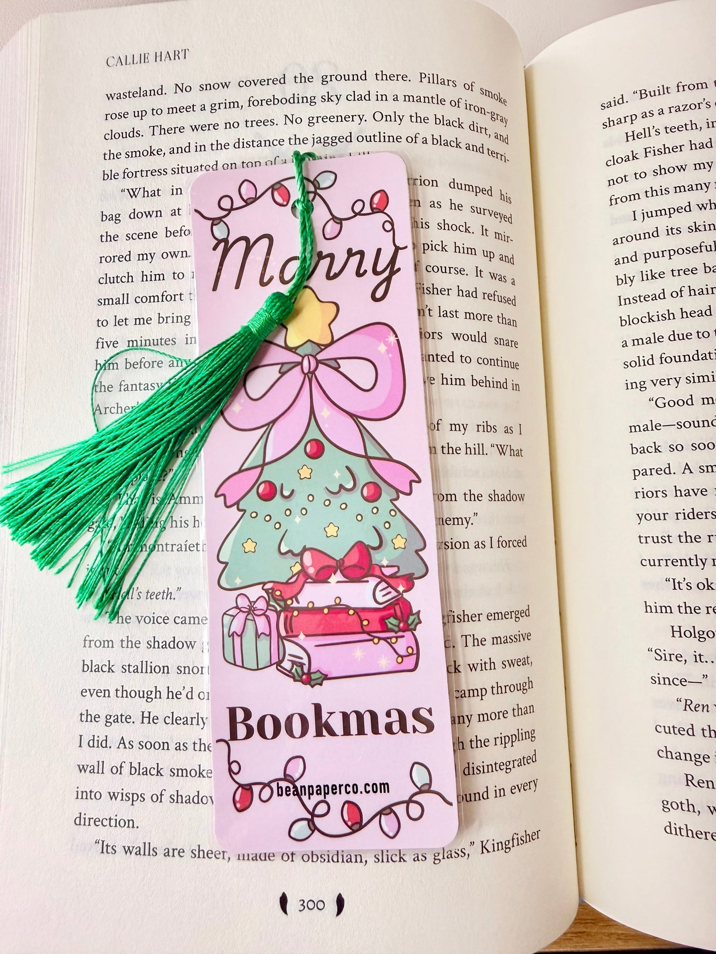 “Pink laminated ‘Merry Bookmas’ bookmark with green tassel featuring a decorated Christmas tree sitting on stacked books inside an open novel.”