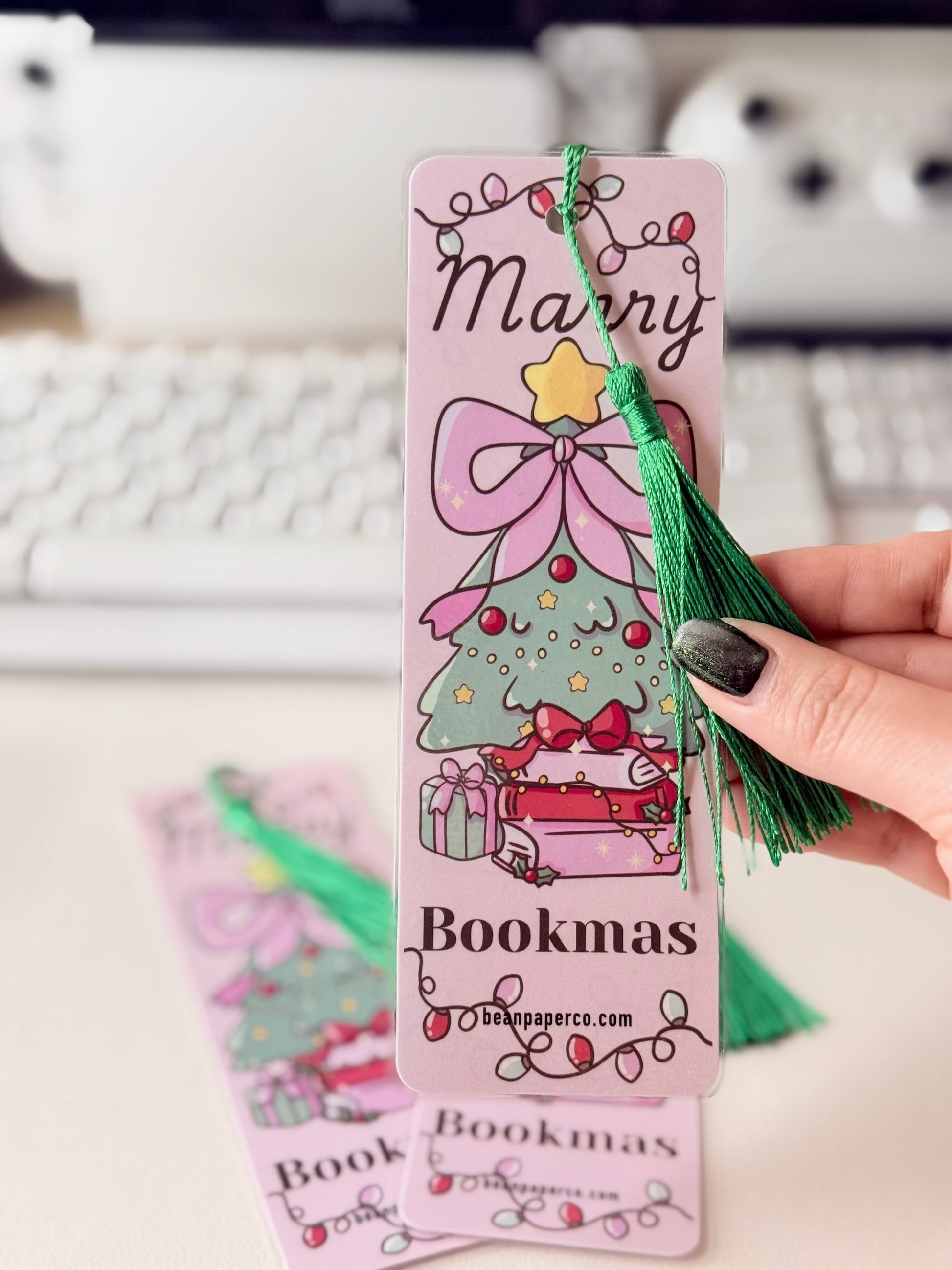 “Close-up of pink Merry Bookmas bookmark with green tassel and illustrated Christmas tree on stacked books.”