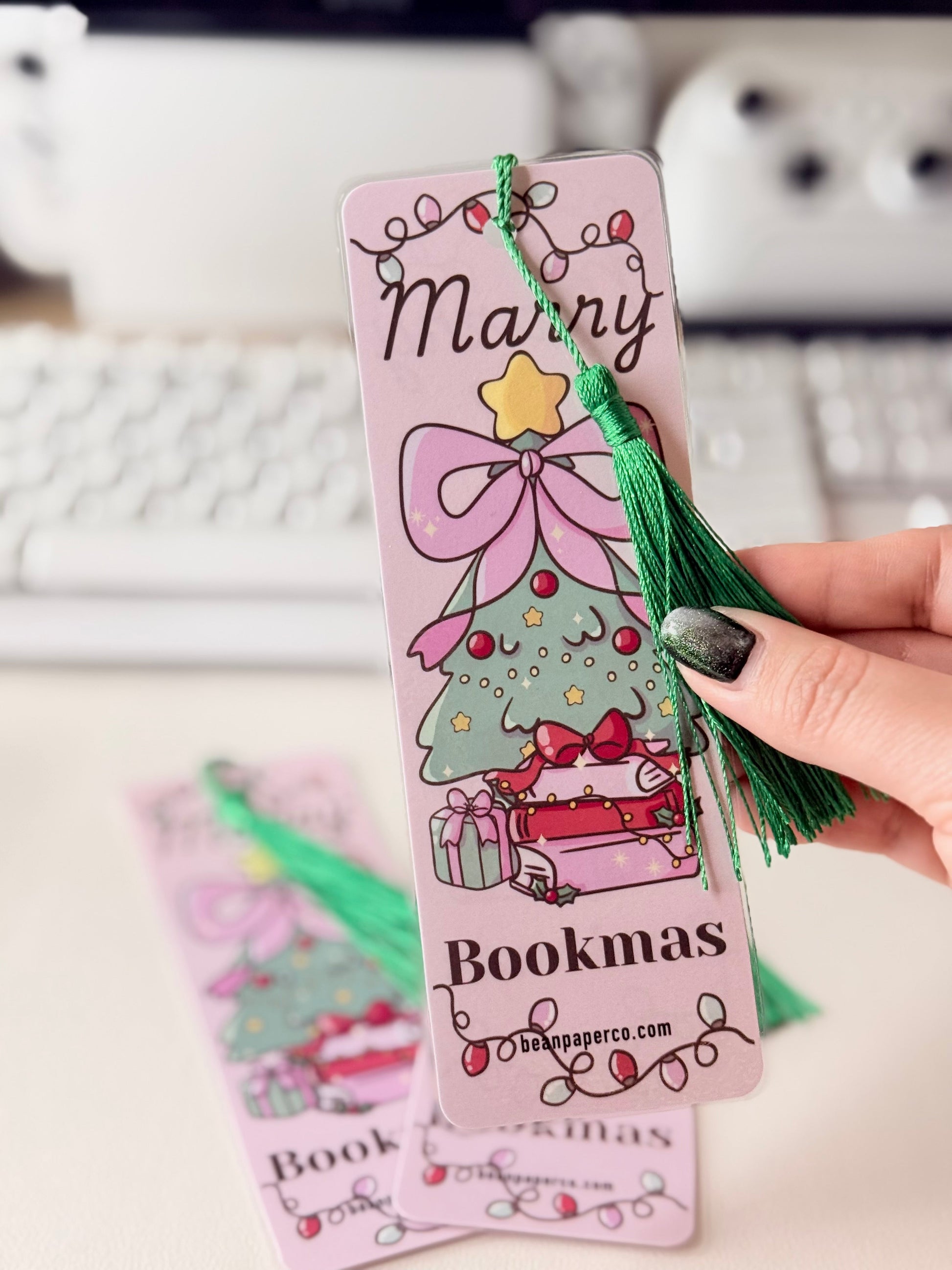 “Close-up of pink Merry Bookmas bookmark with green tassel and illustrated Christmas tree on stacked books.”