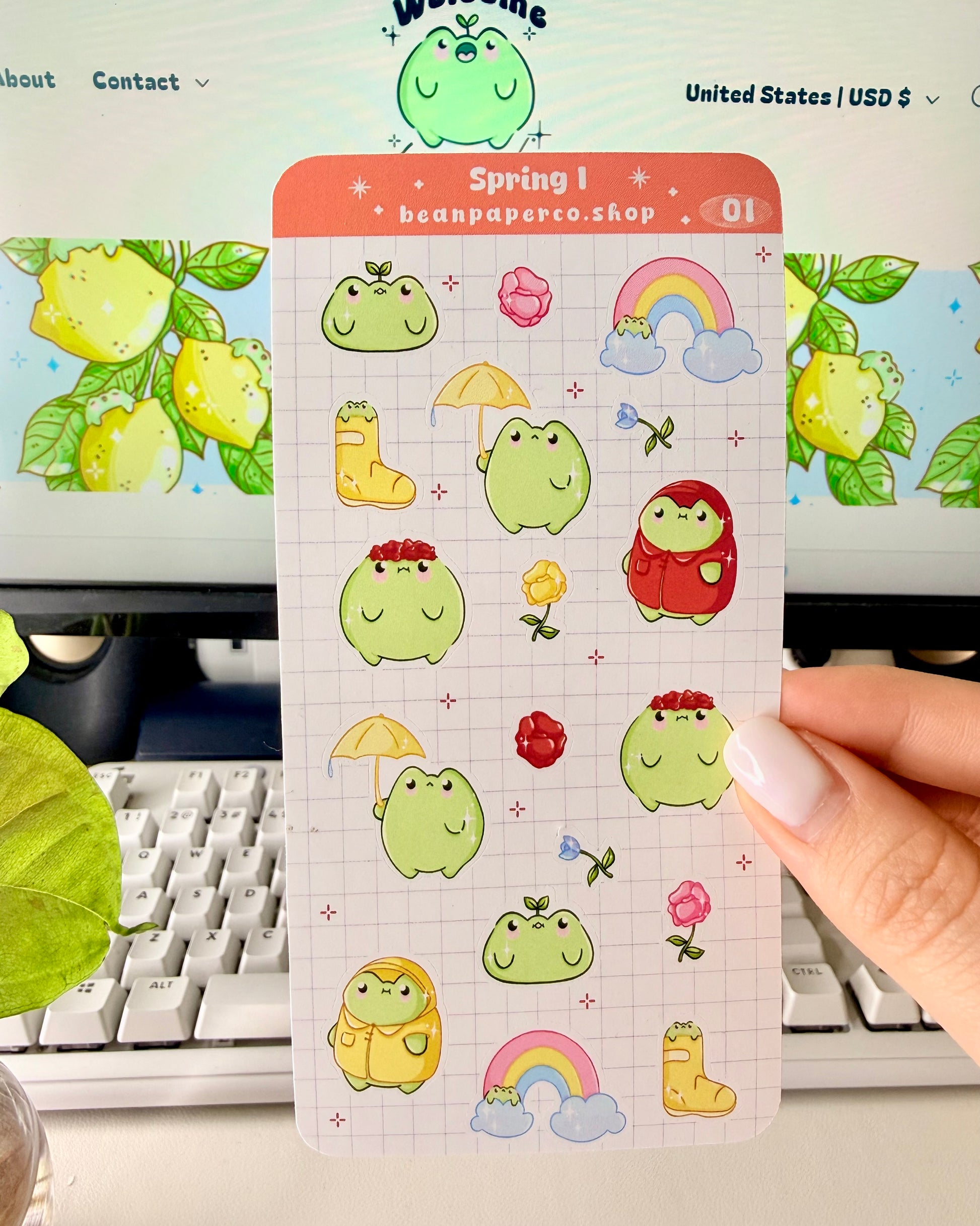 Hand holding a spring-themed sticker sheet with cute green frog characters, flowers, rain boots, umbrellas, rainbows, and potted plants, photographed in front of a computer screen and plant.