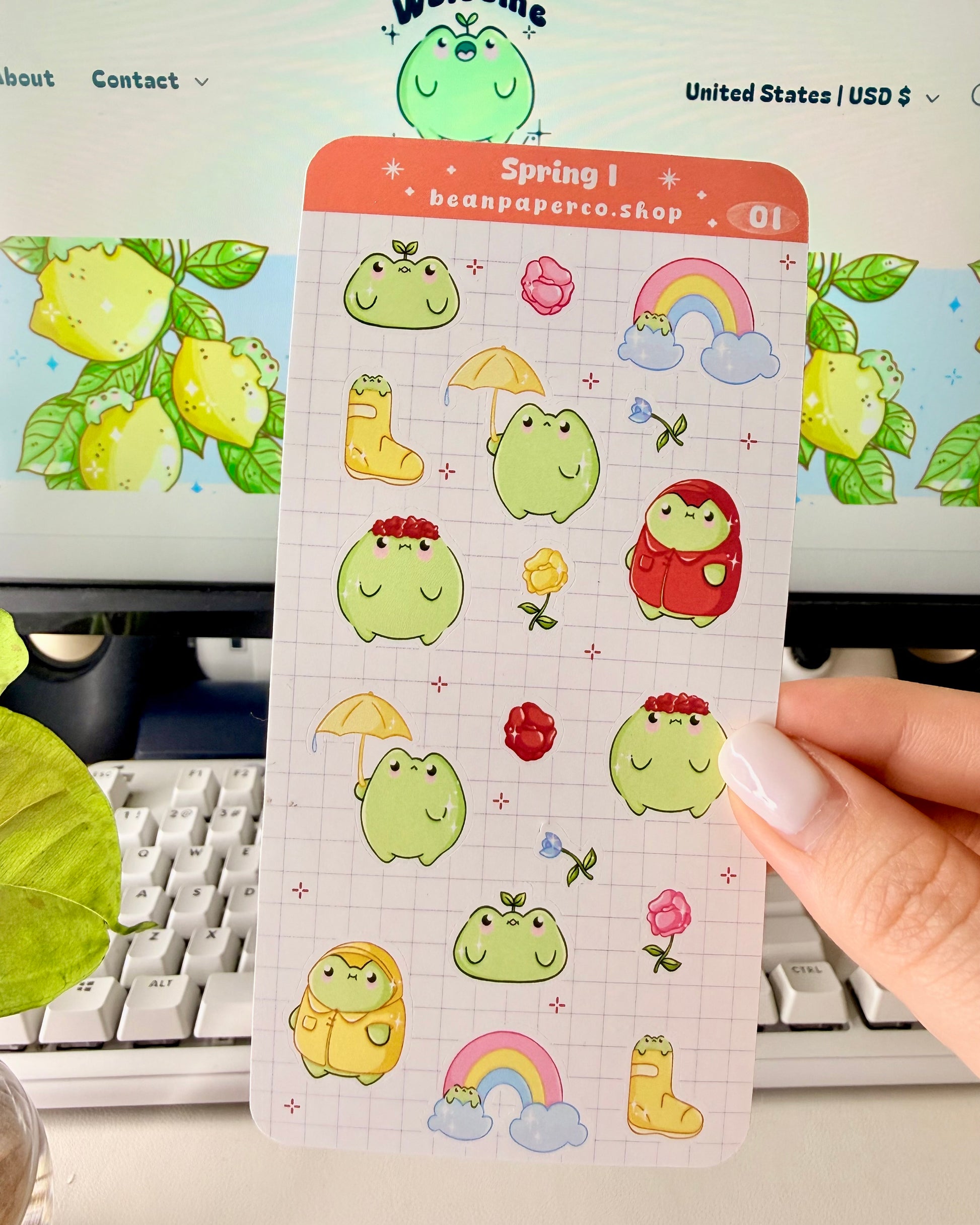 Hand holding a spring-themed sticker sheet with cute green frog characters, flowers, rain boots, umbrellas, rainbows, and potted plants, photographed in front of a computer screen and plant.