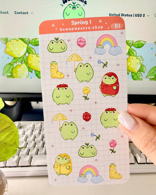 Hand holding a spring-themed sticker sheet with cute green frog characters, flowers, rain boots, umbrellas, rainbows, and potted plants, photographed in front of a computer screen and plant.