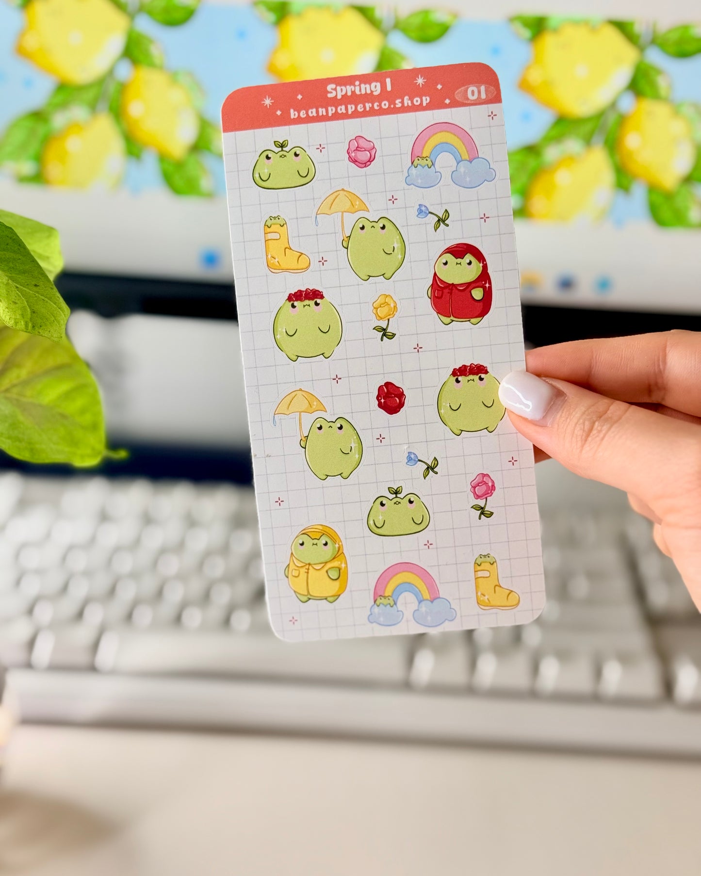 Hand holding a spring-themed sticker sheet with cute green frog characters, flowers, rain boots, umbrellas, rainbows, and potted plants, photographed in front of a computer screen and plant.