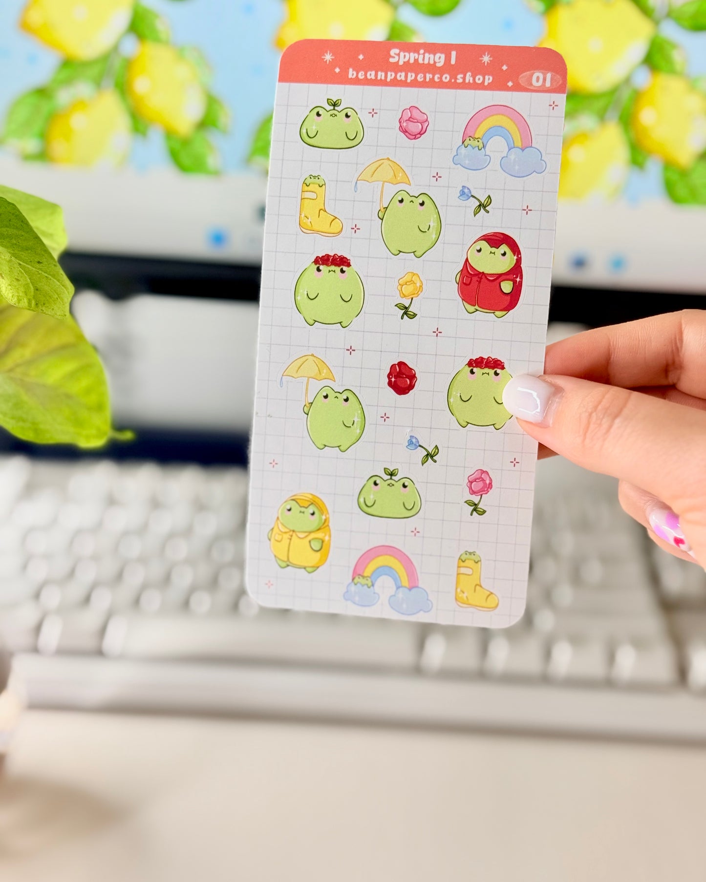 Hand holding a spring-themed sticker sheet with cute green frog characters, flowers, rain boots, umbrellas, rainbows, and potted plants, photographed in front of a computer screen and plant.