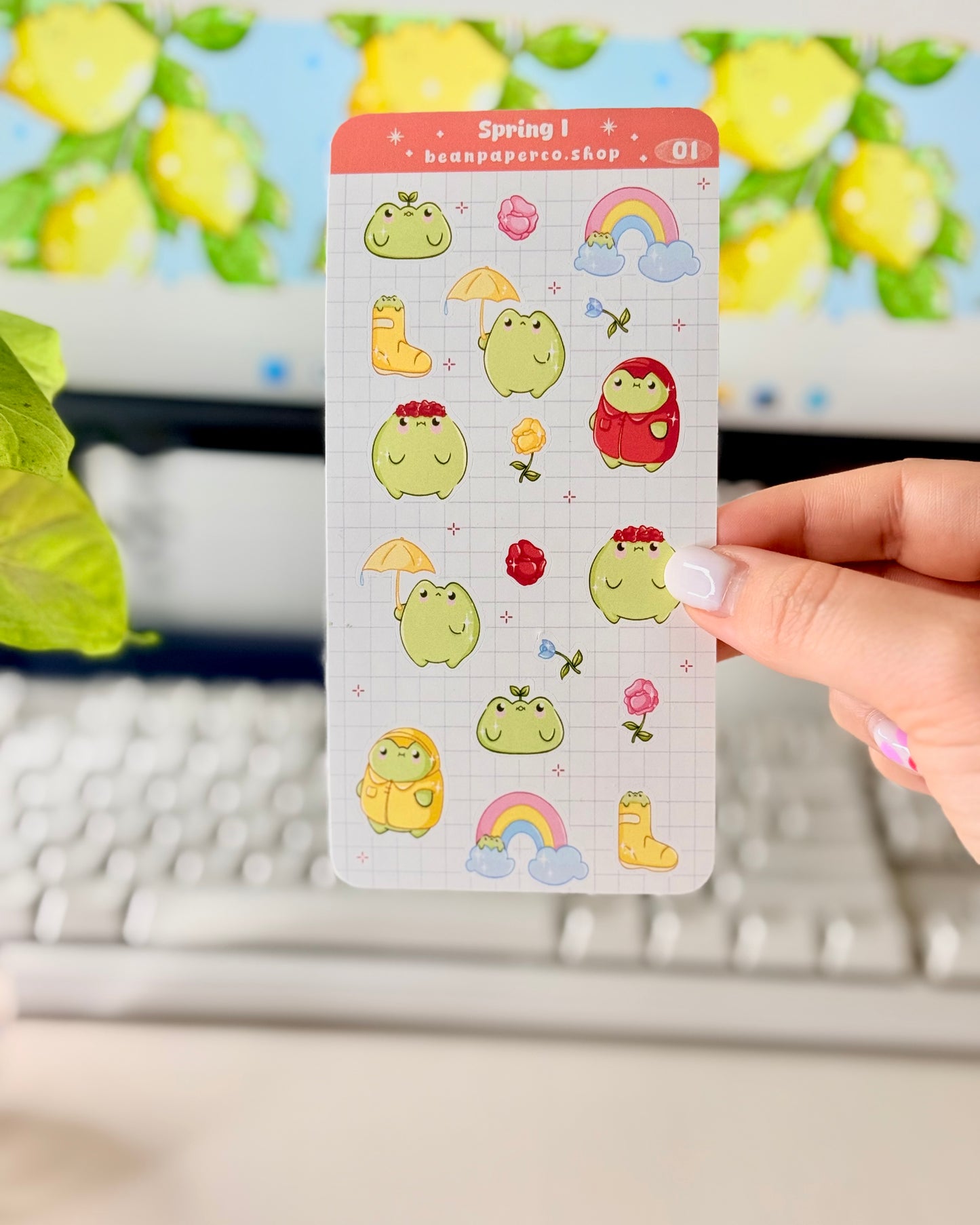 Hand holding a spring-themed sticker sheet with cute green frog characters, flowers, rain boots, umbrellas, rainbows, and potted plants, photographed in front of a computer screen and plant.