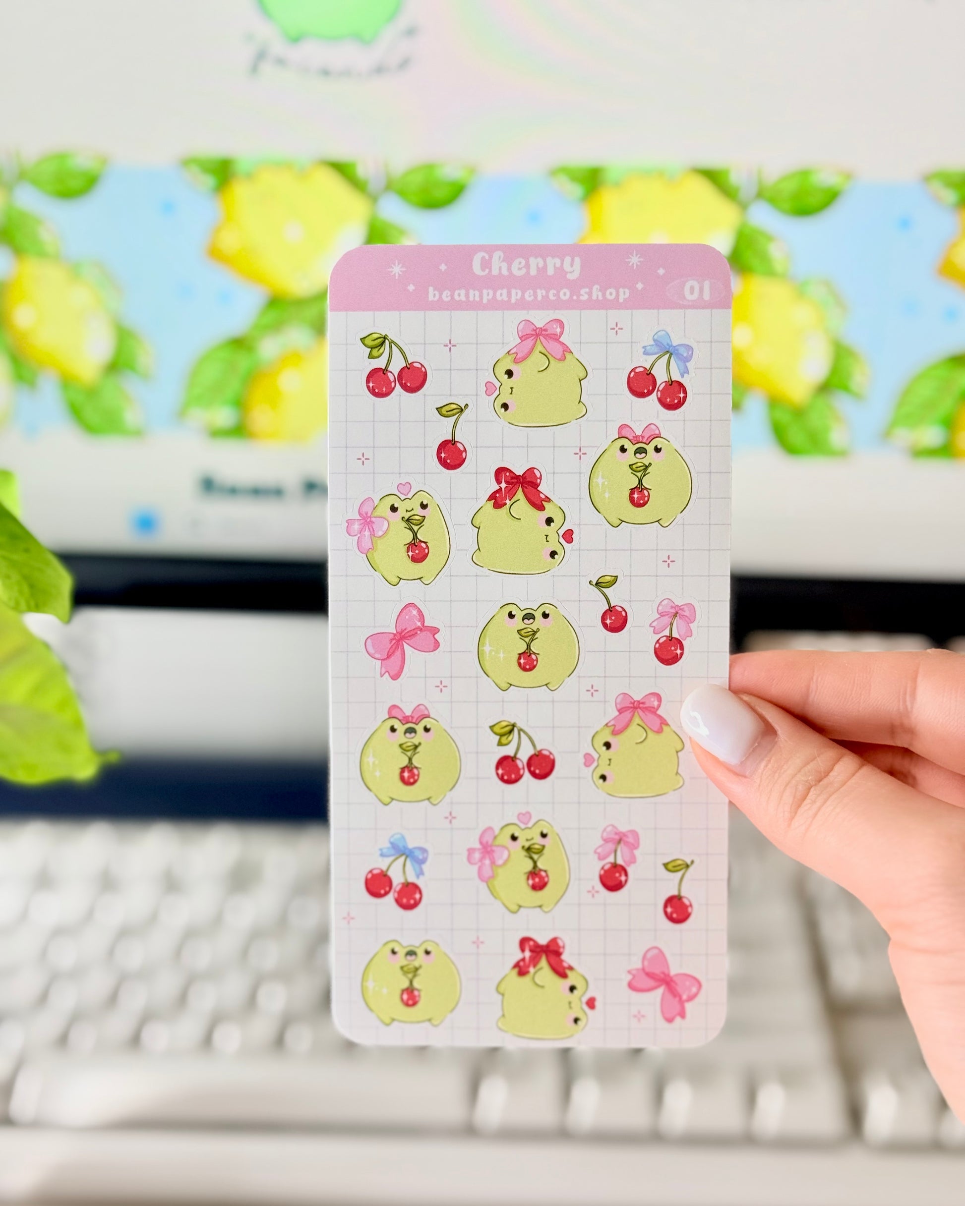 Hand holding a Cherry-themed frog sticker sheet featuring cute green frogs with pink bows, and red cherries.