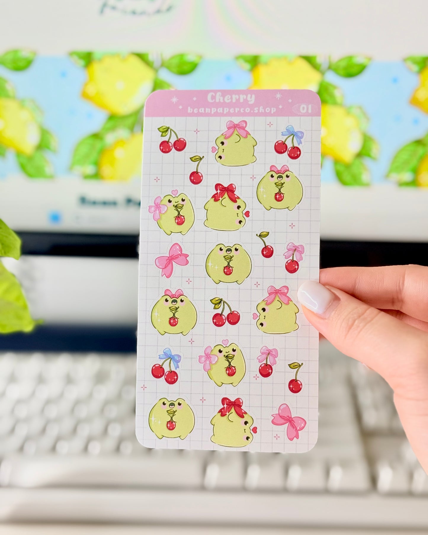 Hand holding a Cherry-themed frog sticker sheet featuring cute green frogs with pink bows, and red cherries.