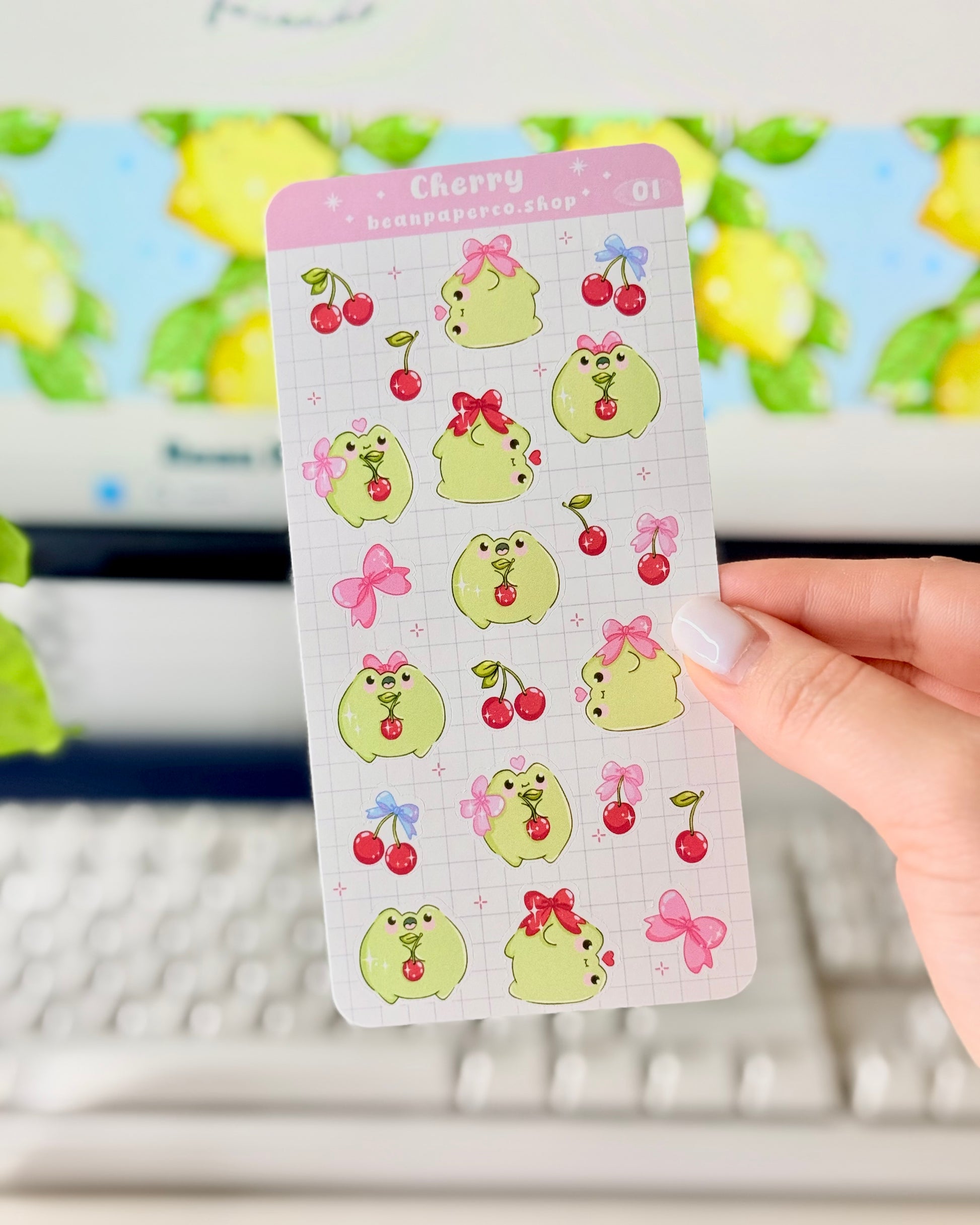 Hand holding a Cherry-themed frog sticker sheet featuring cute green frogs with pink bows, and red cherries.