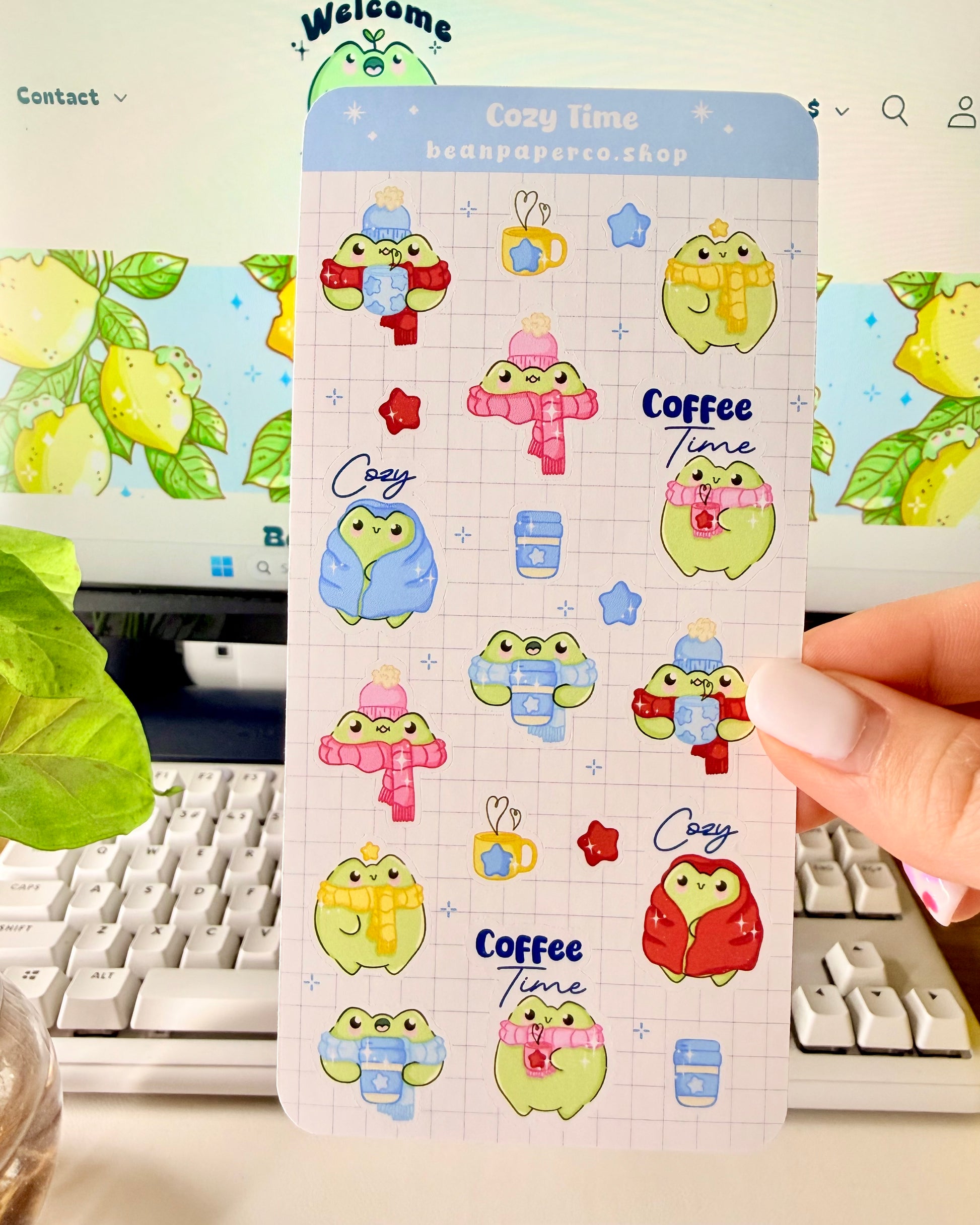 Hand holding a cozy frog sticker sheet featuring cute green frogs in scarves and hats, holding coffee cups, and wrapped in blankets.