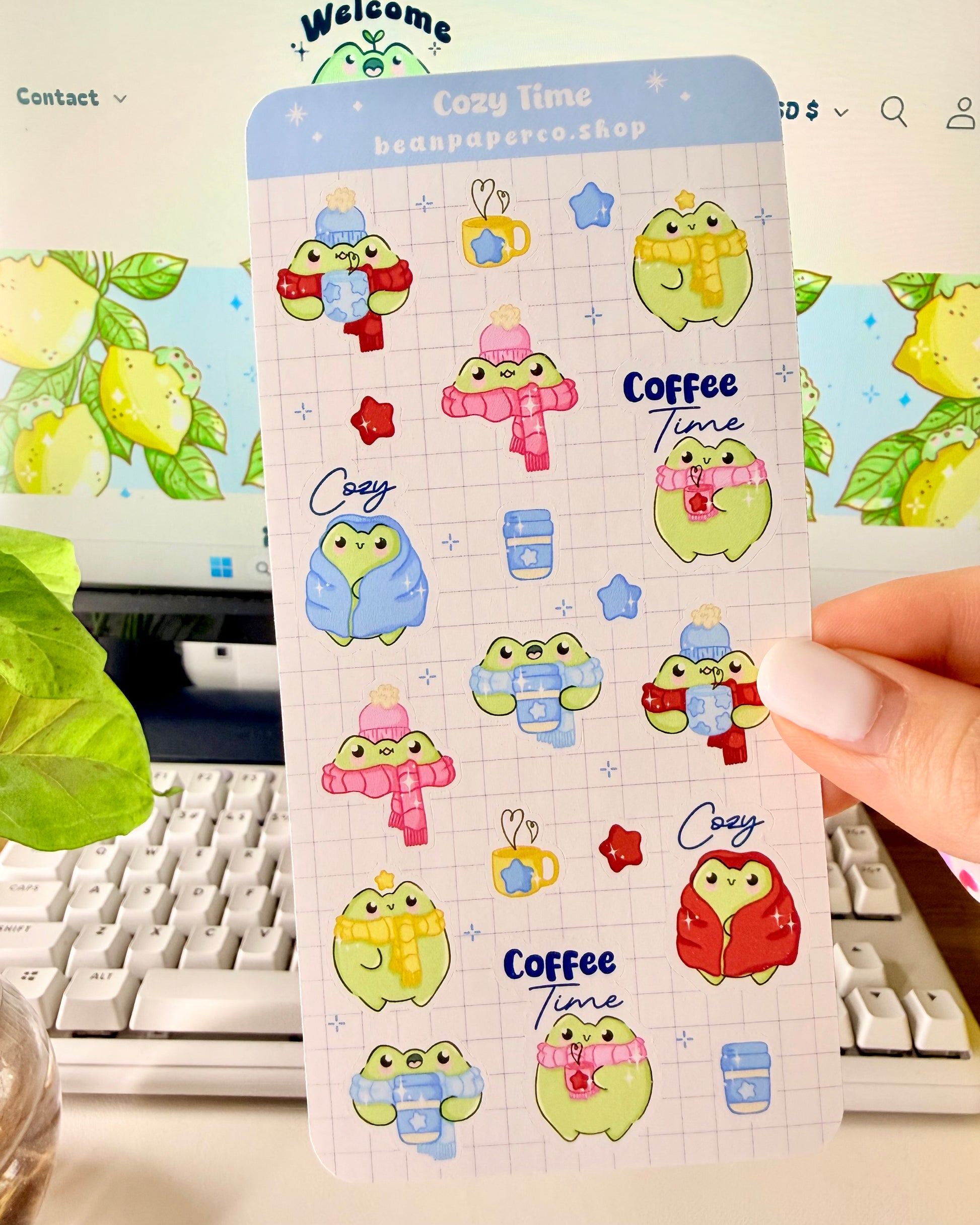 Hand holding a cozy frog sticker sheet featuring cute green frogs in scarves and hats, holding coffee cups, and wrapped in blankets.